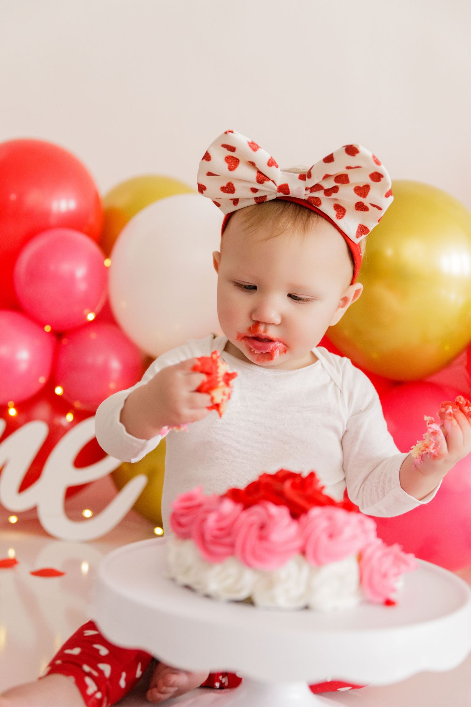Valentine's themed cake smash setup with pink and gold balloons and a small white cake.