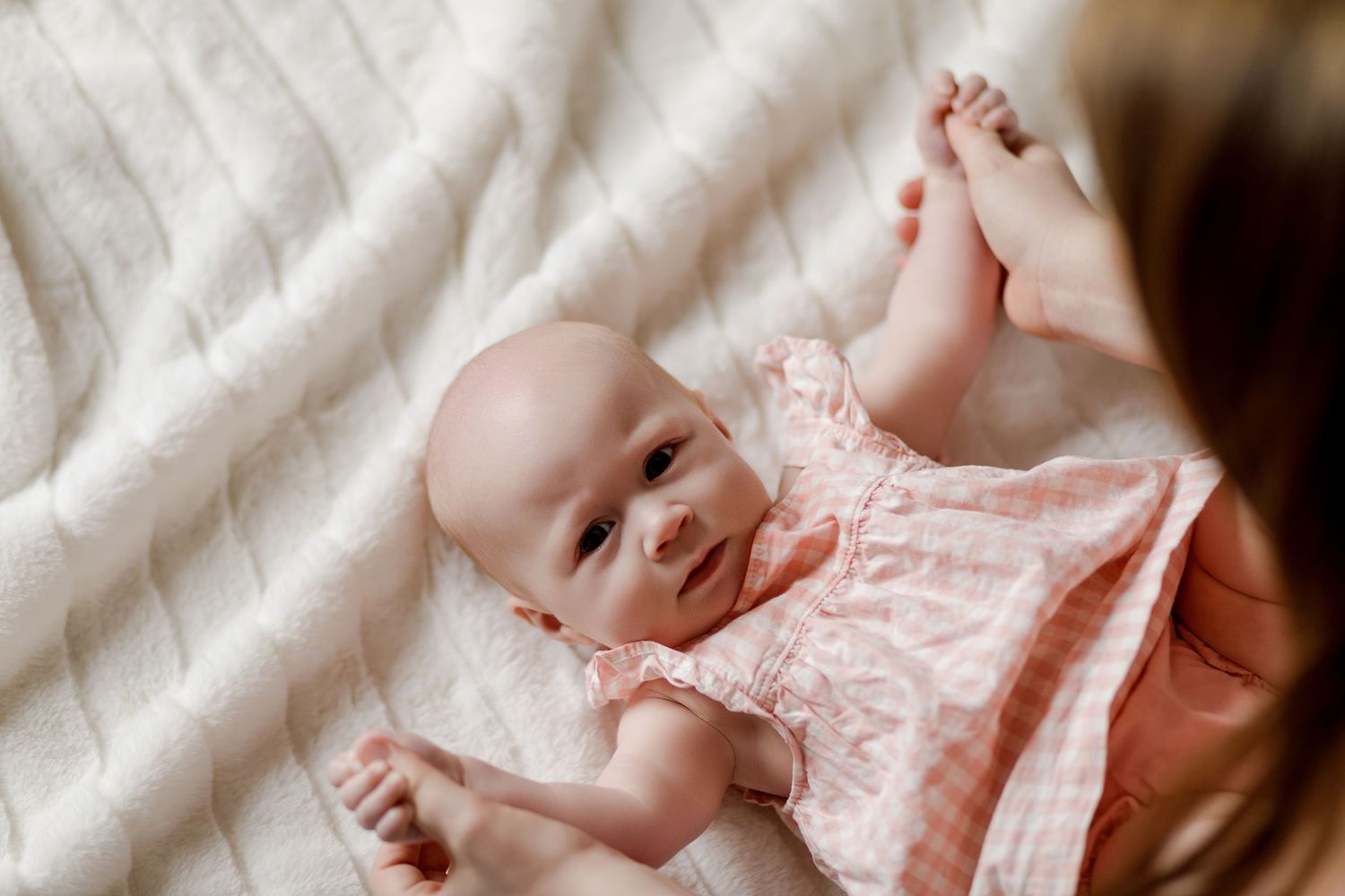 Adorable baby girl in a pink ruffled dress lies on a white blanket during a lifestyle photo session.