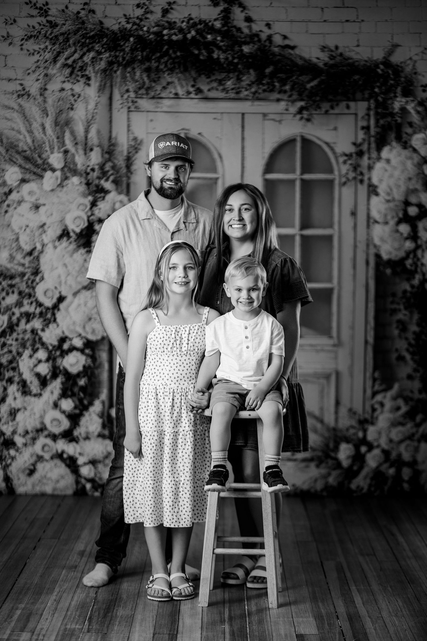 Black and white family portrait of parents with two young children posed in front of floral backdrop with vintage door.