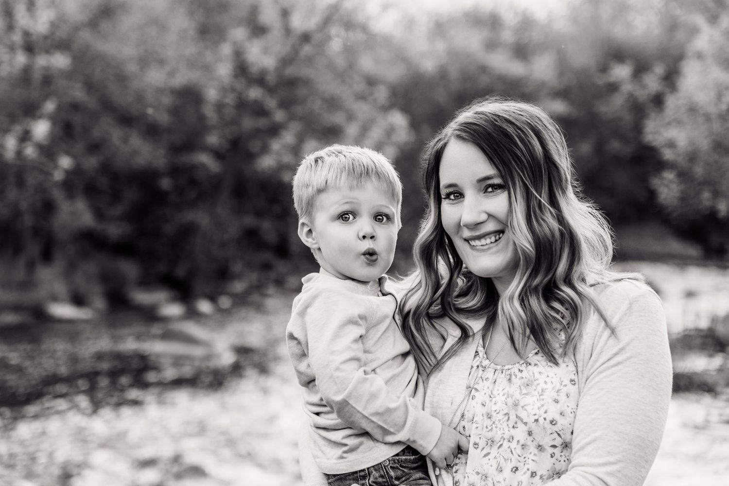 Black and white photo of a smiling woman holding a young toddler boy outdoors near trees.