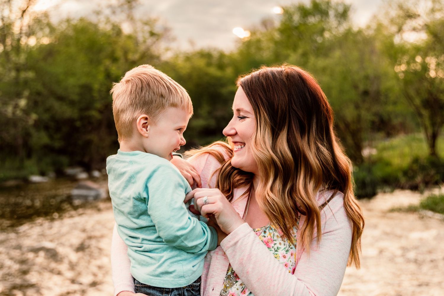 A mother holds her young toddler boy outdoors near a creek, sharing a sweet smiling moment together.