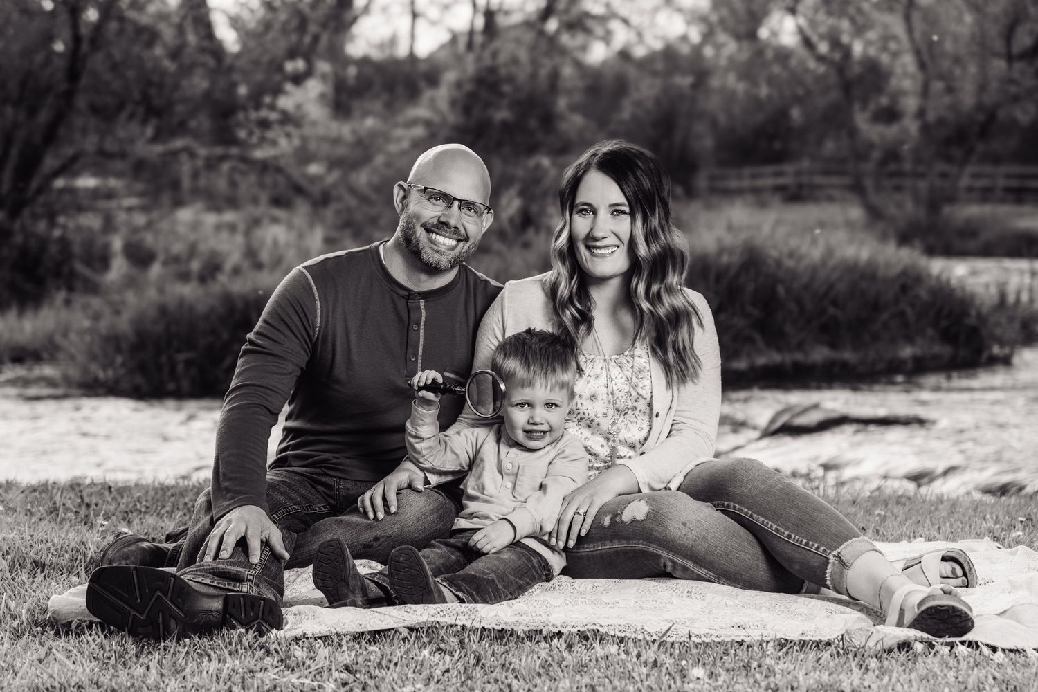Black and white family portrait of smiling parents with young child sitting on blanket outdoors in park setting.