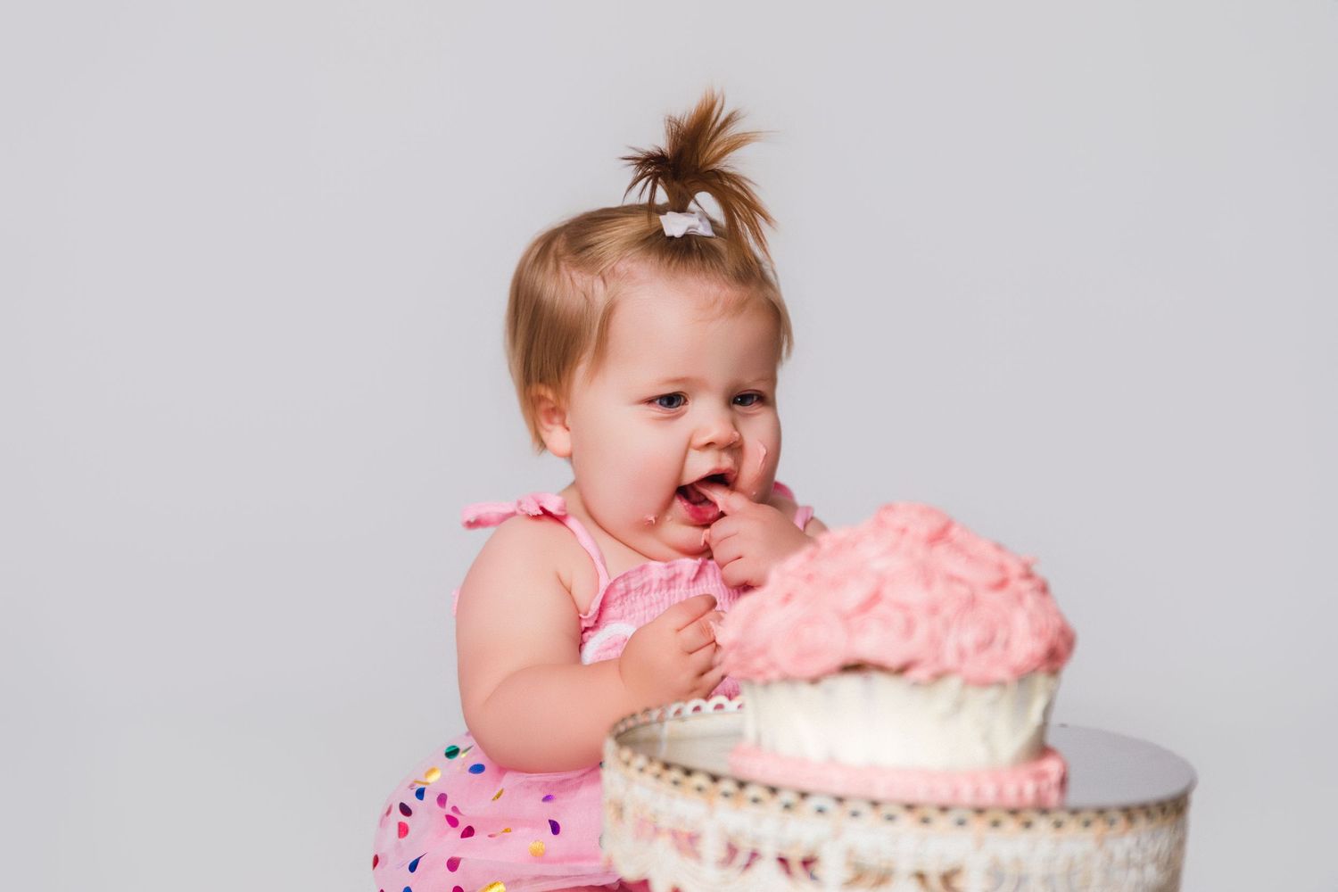 Baby in pink outfit enjoying cake smash photo session with decorated white cake stand.