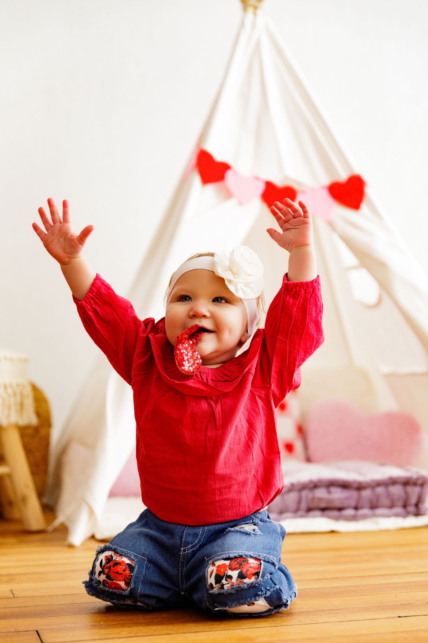 Baby in red sweater and bow raises arms joyfully in front of teepee with heart decorations.
