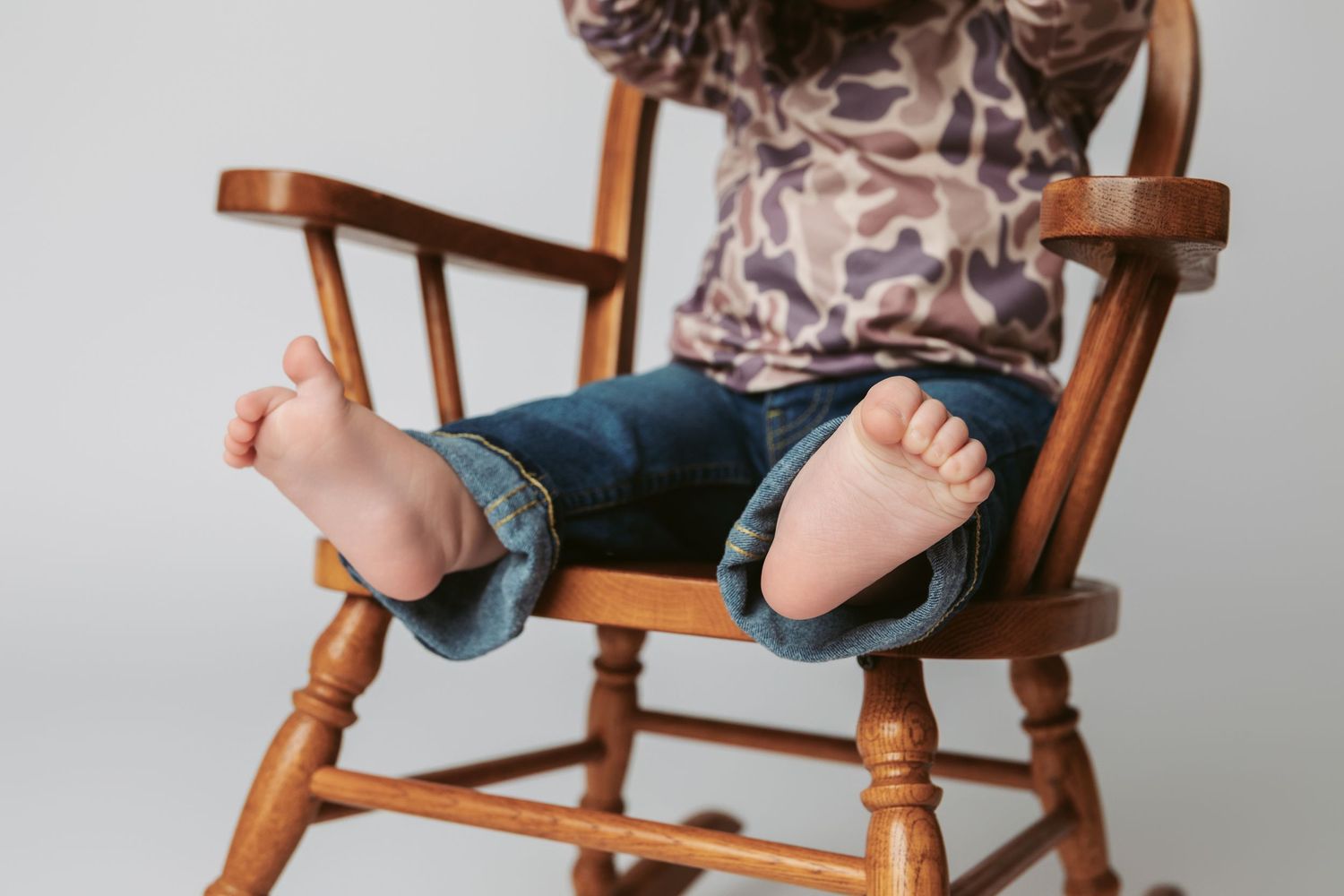 Small feet in blue jeans dangle from a wooden rocking chair against a white background.