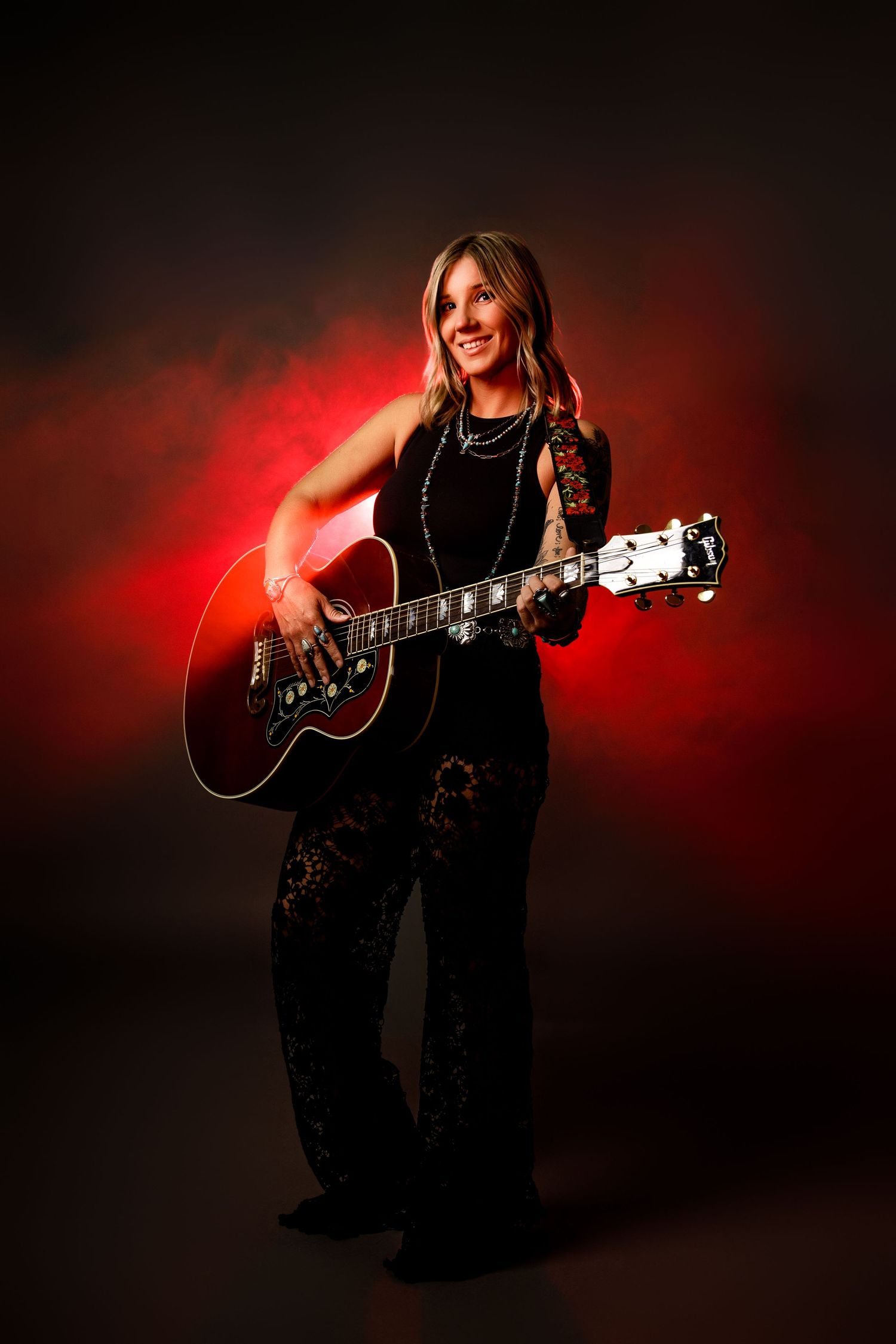 Woman in black outfit playing acoustic guitar against dark background with red smoke lighting.