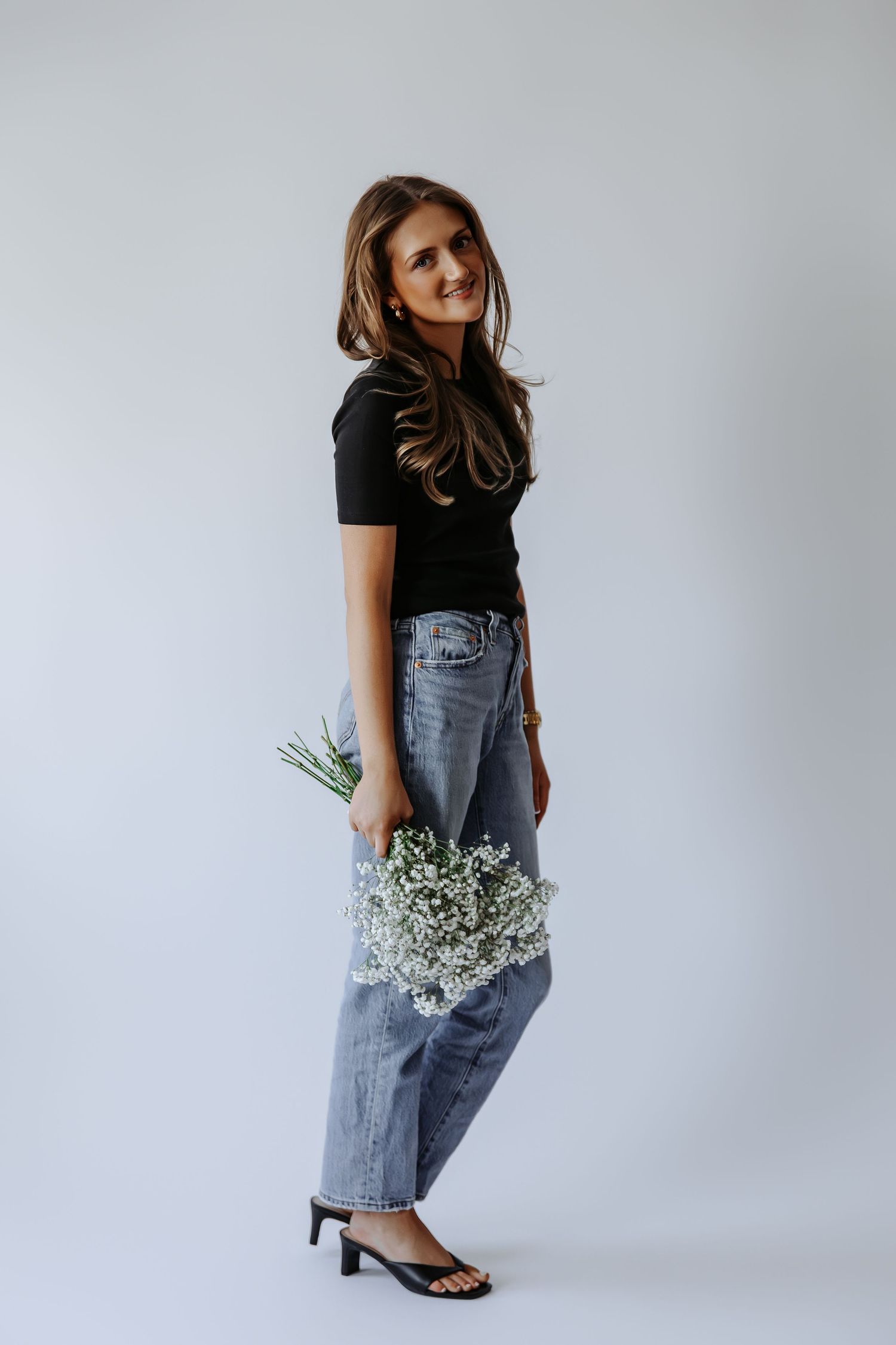 Smiling woman in black top and jeans holds white baby's breath flowers in bright studio setting.
