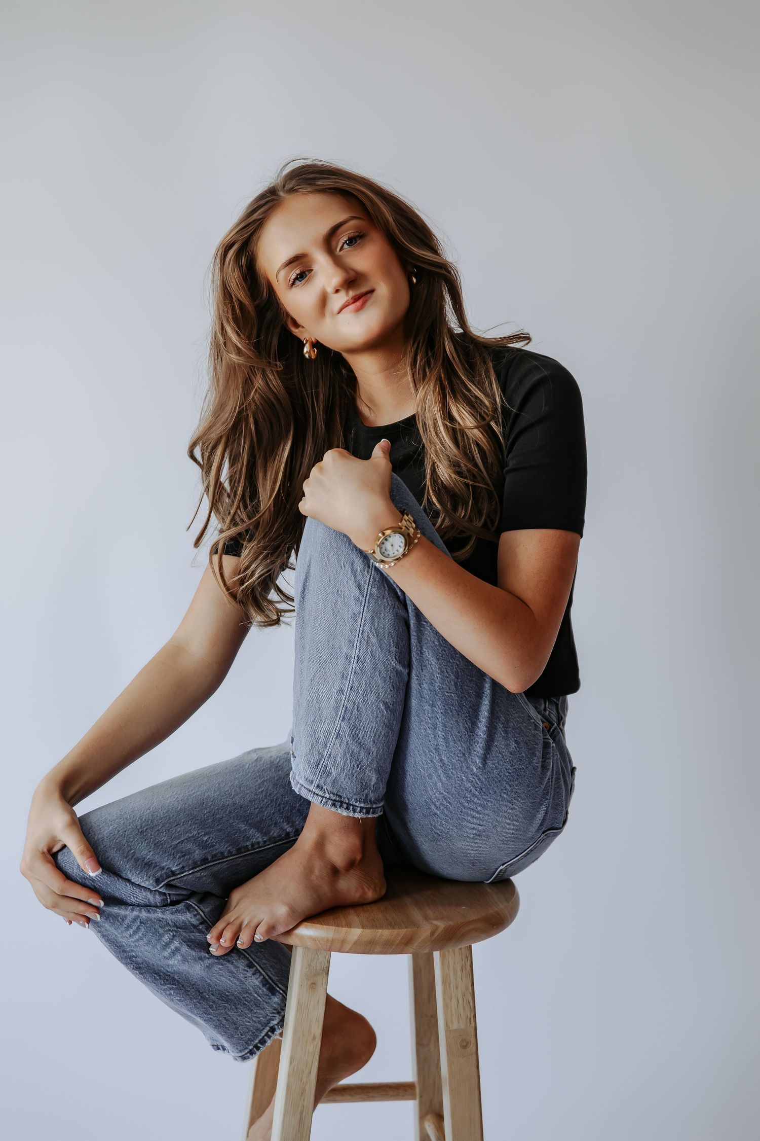 Young woman with long wavy hair sits barefoot on wooden stool wearing black tee and jeans, smiling at camera.