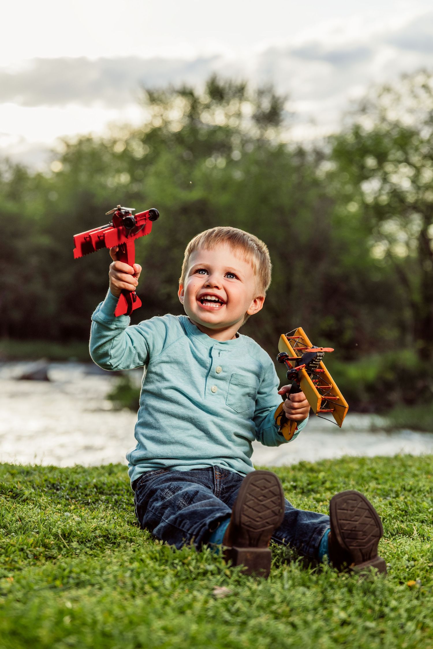 Happy toddler boy sitting on green grass outdoors, joyfully holding red and yellow toy construction vehicles.