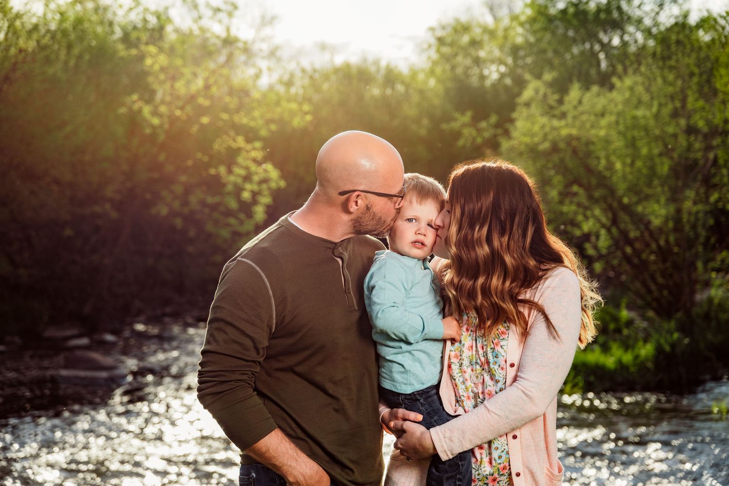 A couple kisses their toddler outdoors by a sunlit creek surrounded by lush green trees.
