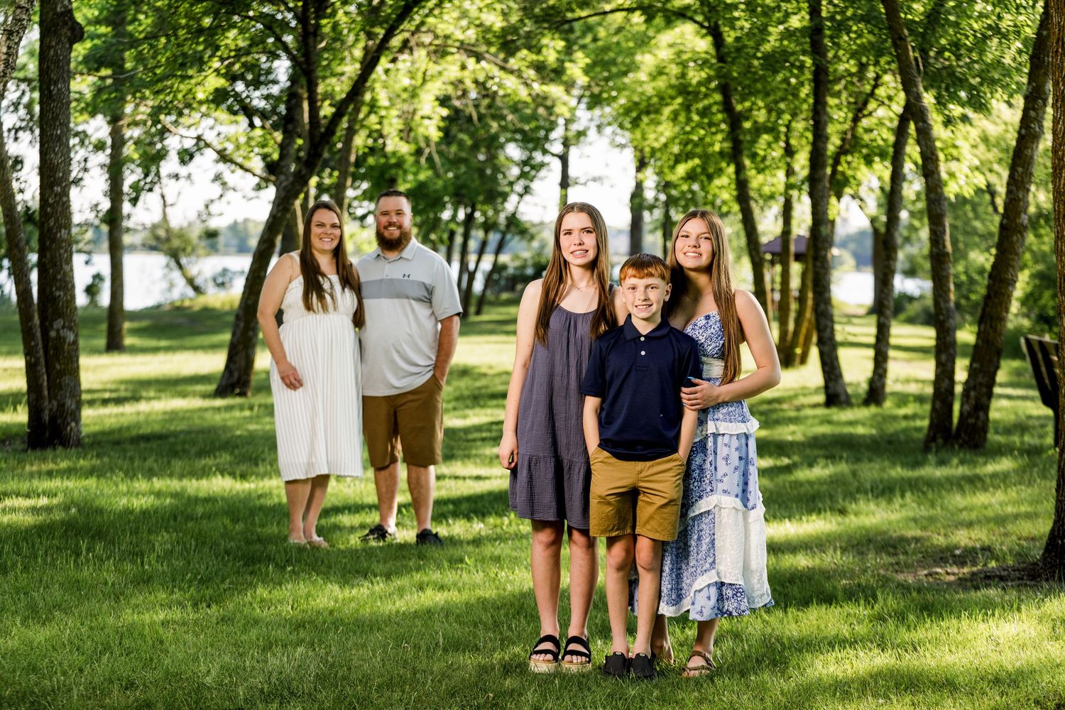 Family of five posing outdoors in a lush green park with trees and a lake visible in the background.