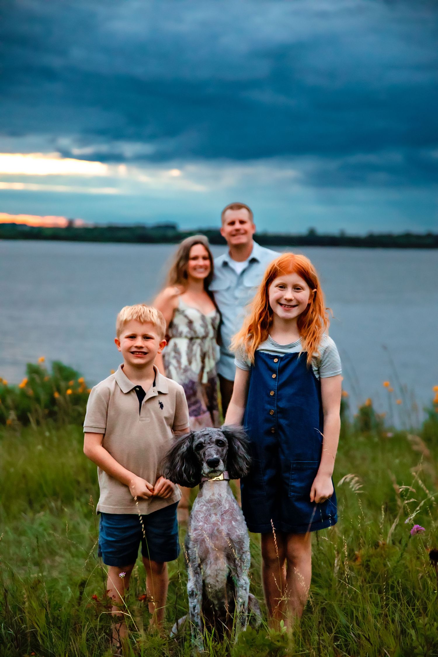 Family with dog poses together in a grassy field by the water under stormy summer skies.