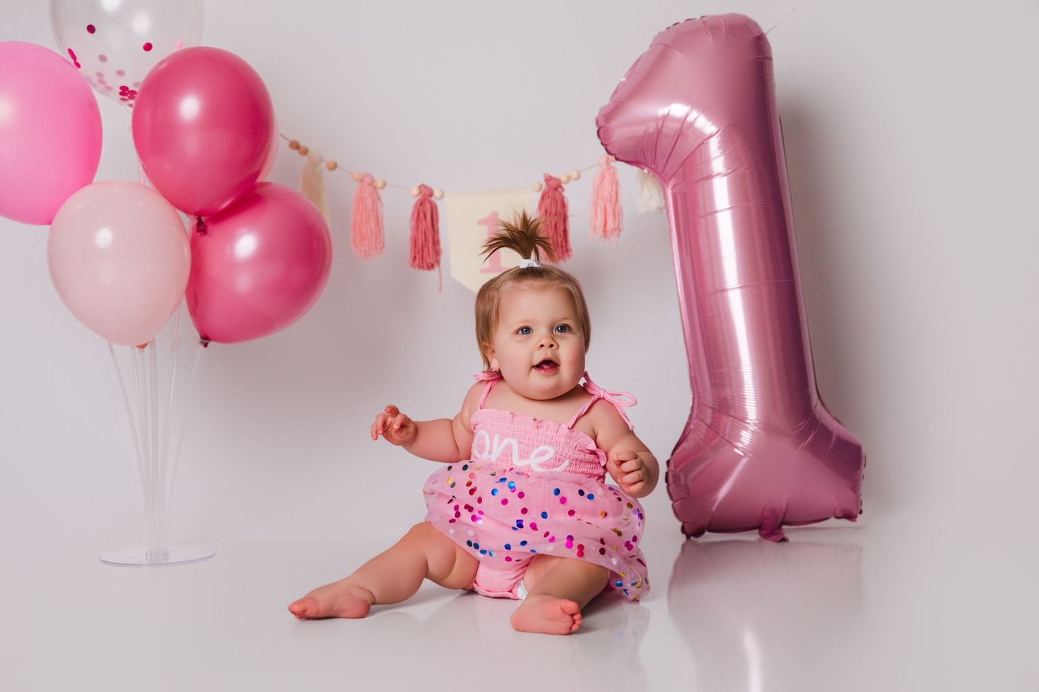 Baby in pink outfit celebrates first birthday with balloons and decorations.