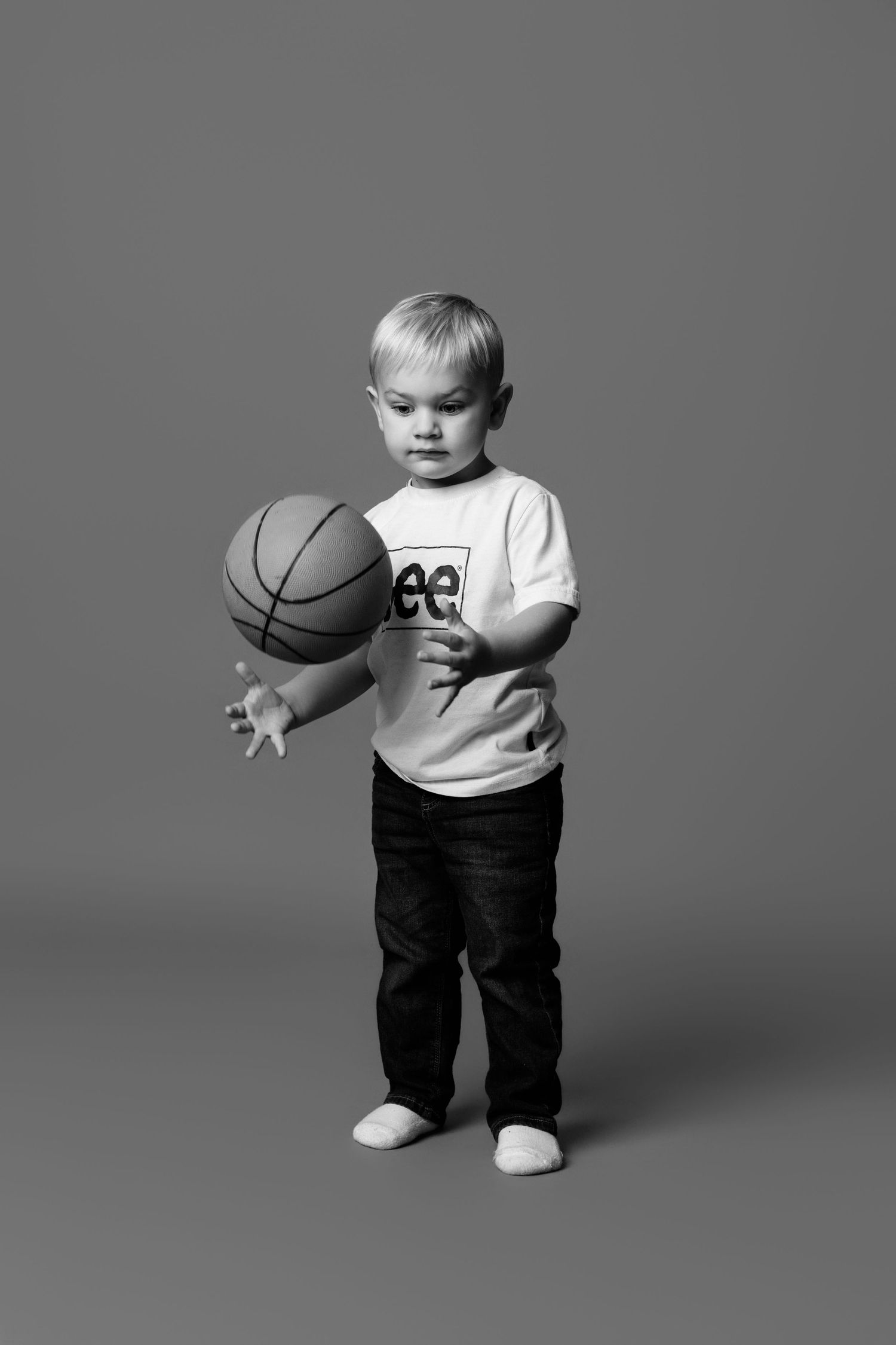 Toddler in white shirt and dark pants playing with basketball in studio setting.