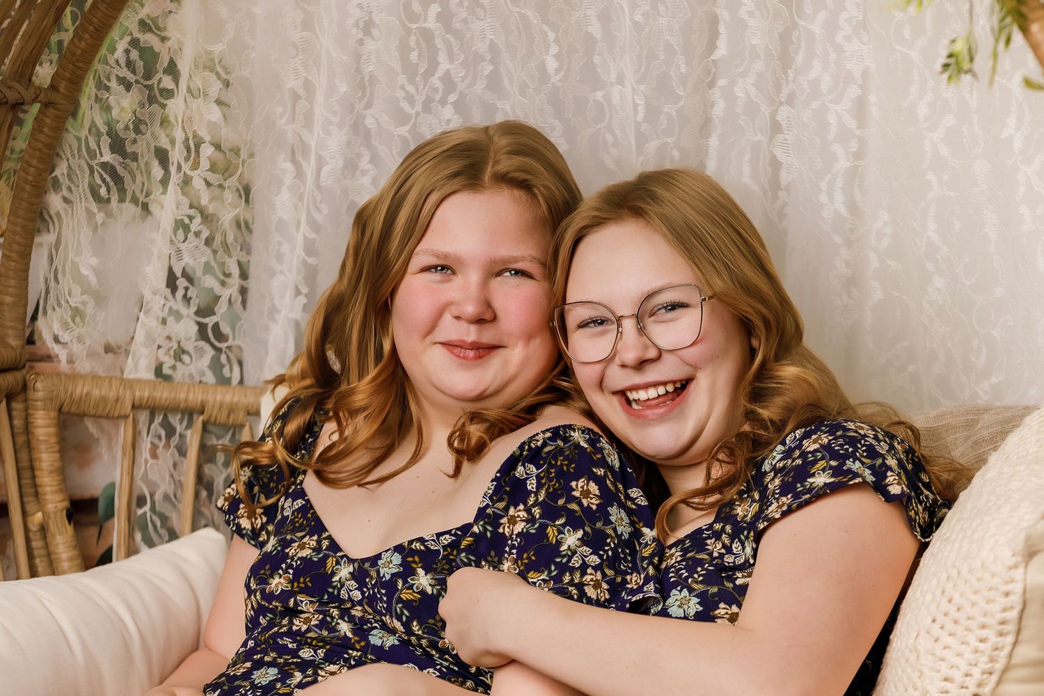 Two young women in matching floral dresses smile and embrace on a cozy settee with lace backdrop.
