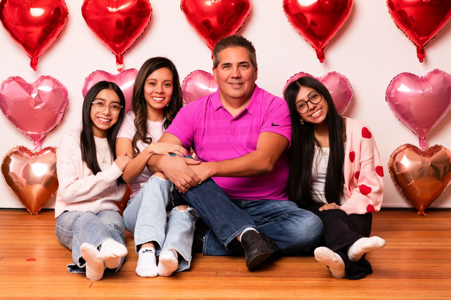 Family of four posing with red and pink heart balloons on Valentine's Day, sitting on wooden floor against white wall.