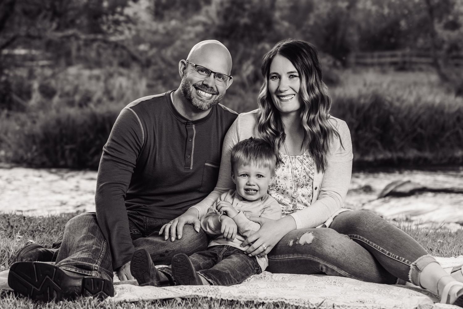Black and white family portrait of smiling couple with toddler sitting on blanket outdoors in nature.