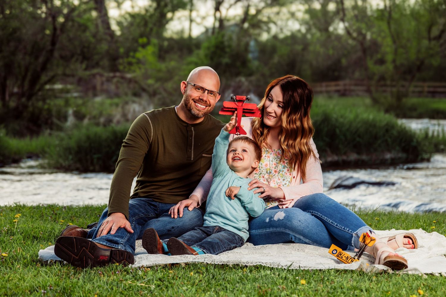 Happy family of three sitting on a blanket by a river, parents smiling with toddler holding a red toy airplane.