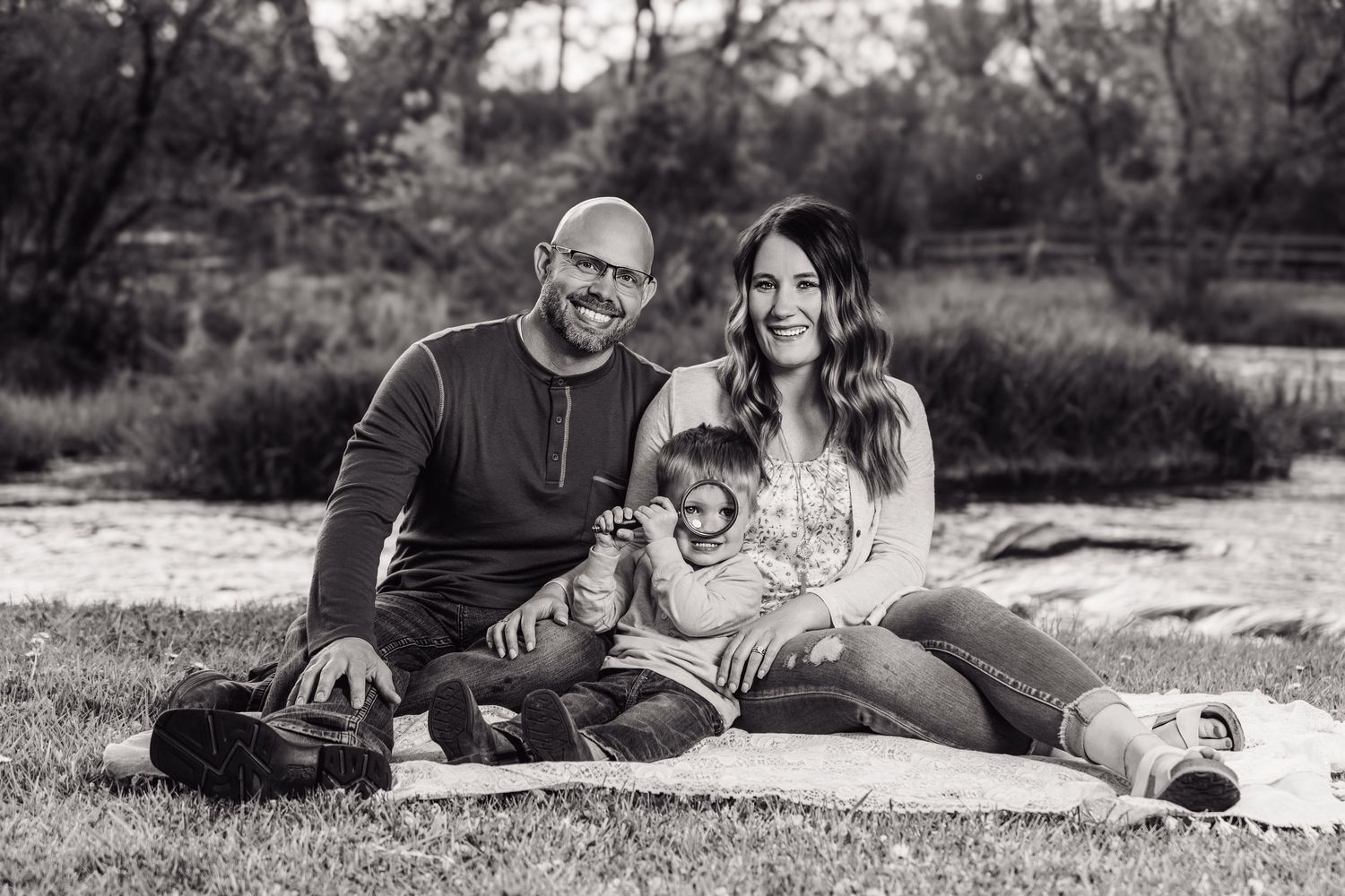 Black and white family portrait of a bald man, woman, and toddler sitting on a blanket outdoors near trees.
