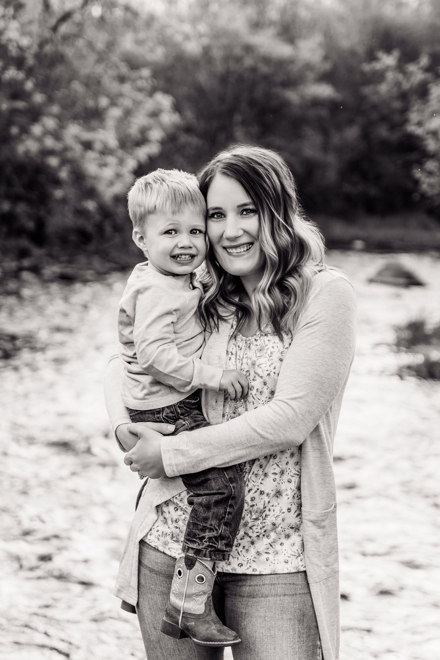 Black and white photo of a smiling woman holding a happy toddler boy outdoors near a creek.