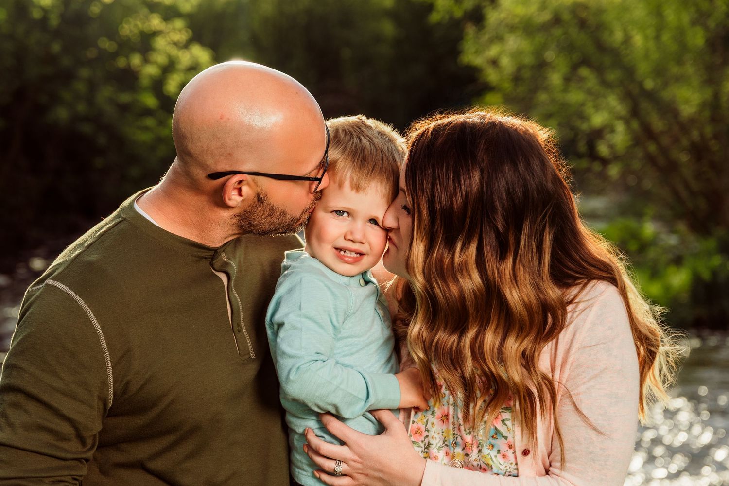 A bald father and brunette mother lovingly kiss their smiling toddler son outdoors in golden sunlight.