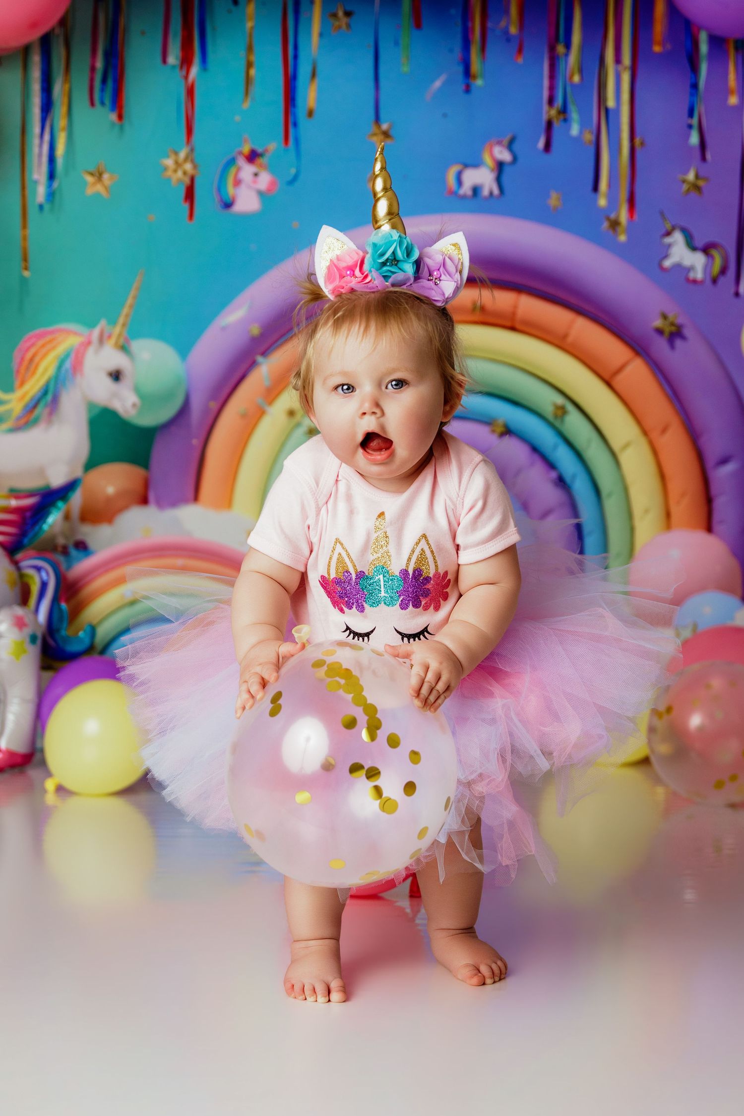 Adorable baby in pink unicorn tutu and gold horn headband at colorful rainbow birthday party.