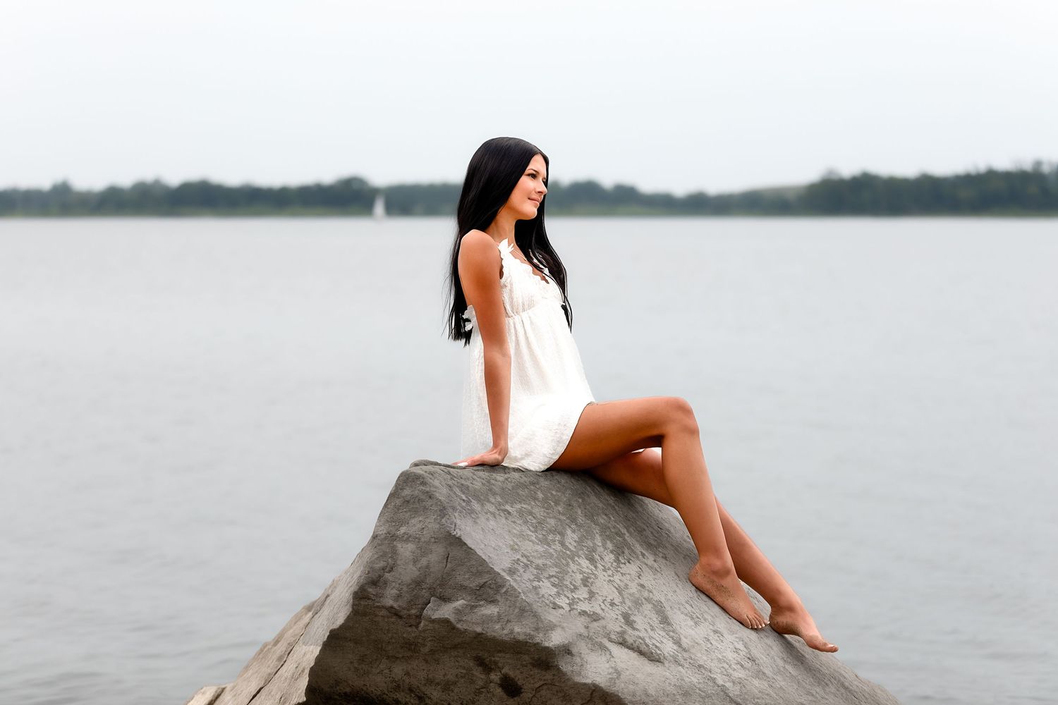 Woman in white swimsuit poses on large rock formation by calm lake on overcast day.
