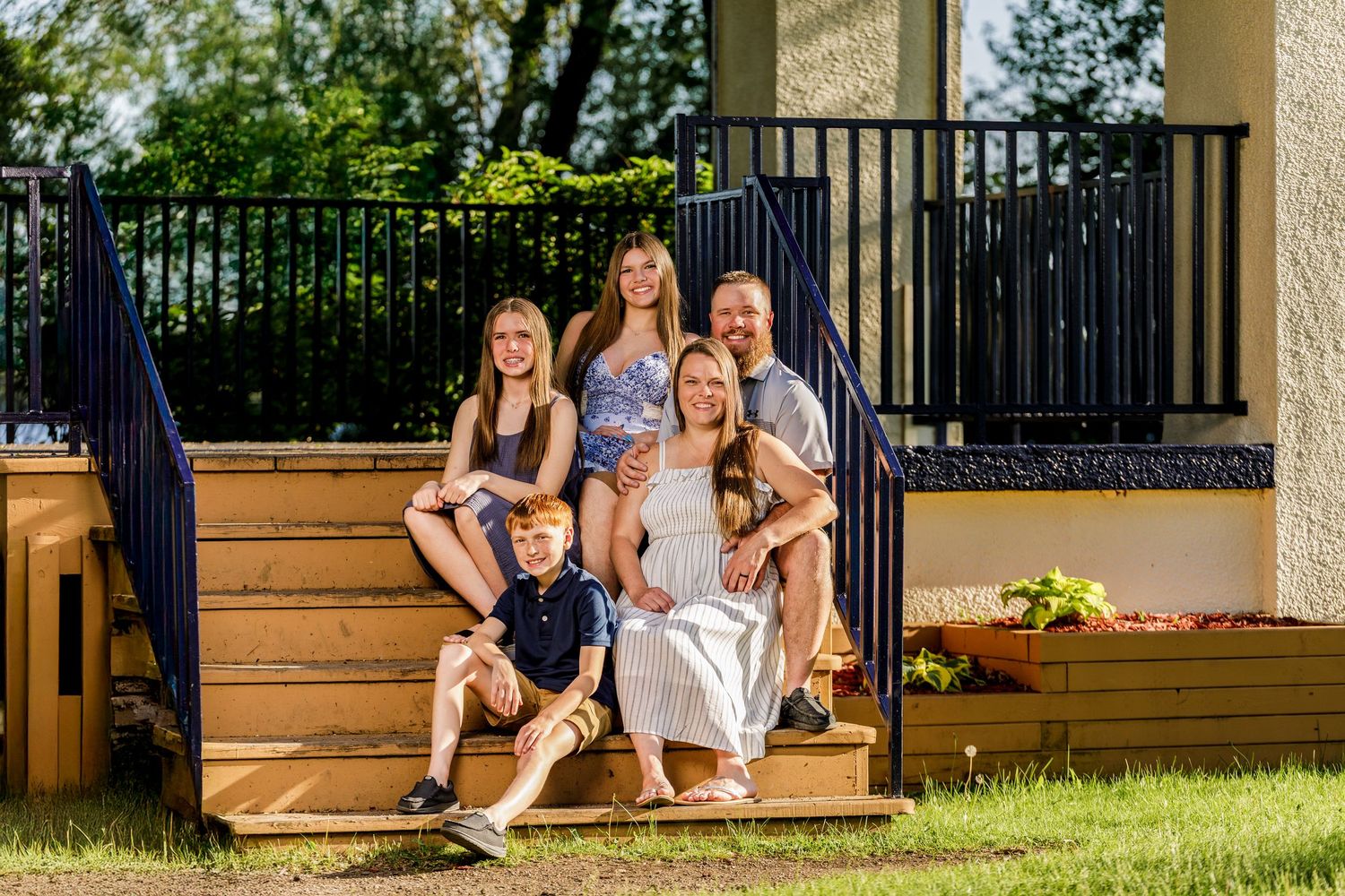 Happy family of five sitting and posing on wooden deck steps in a sunny backyard setting.
