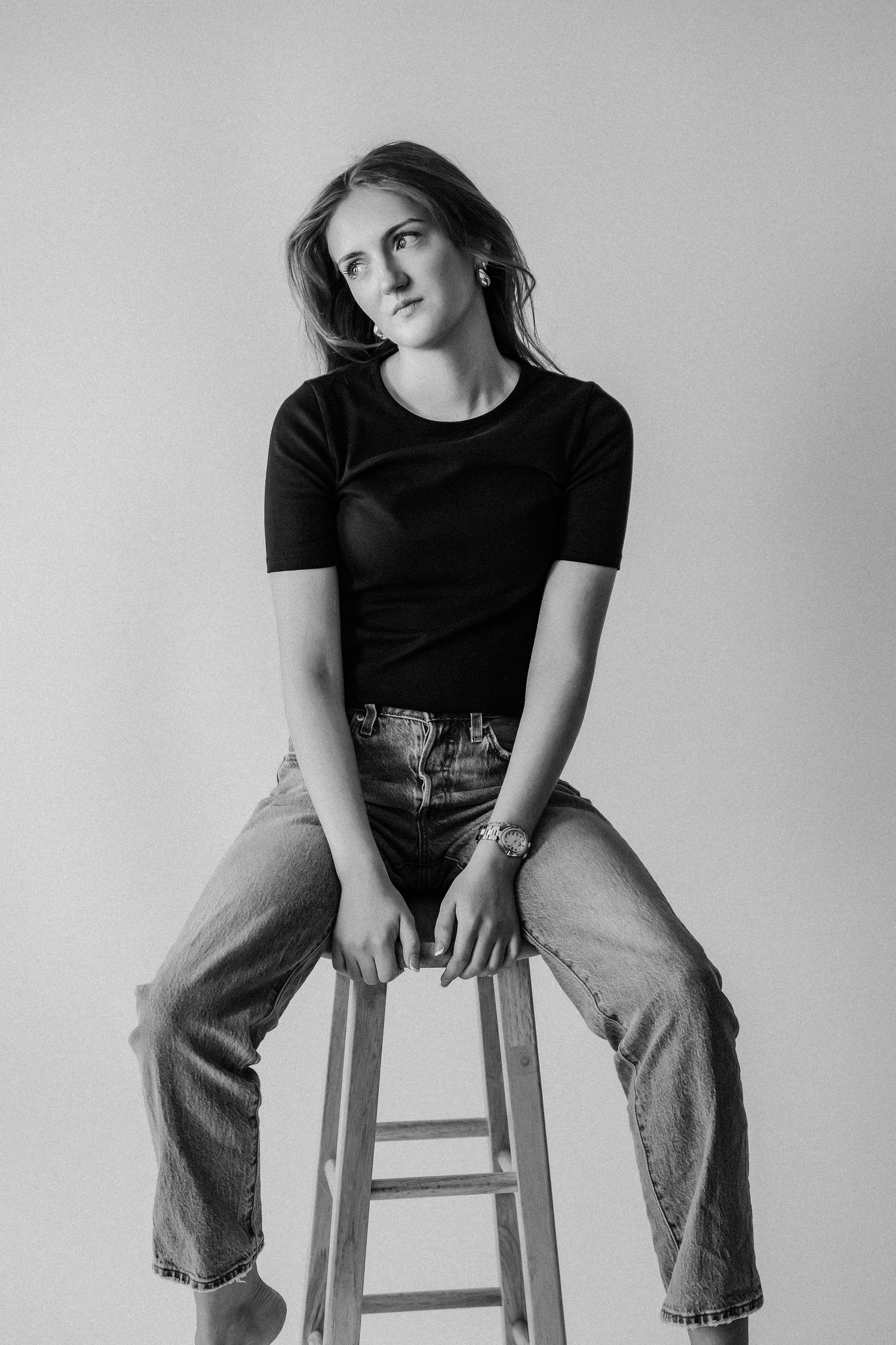 Young woman in black tee and jeans sits on wooden stool in black and white studio portrait.