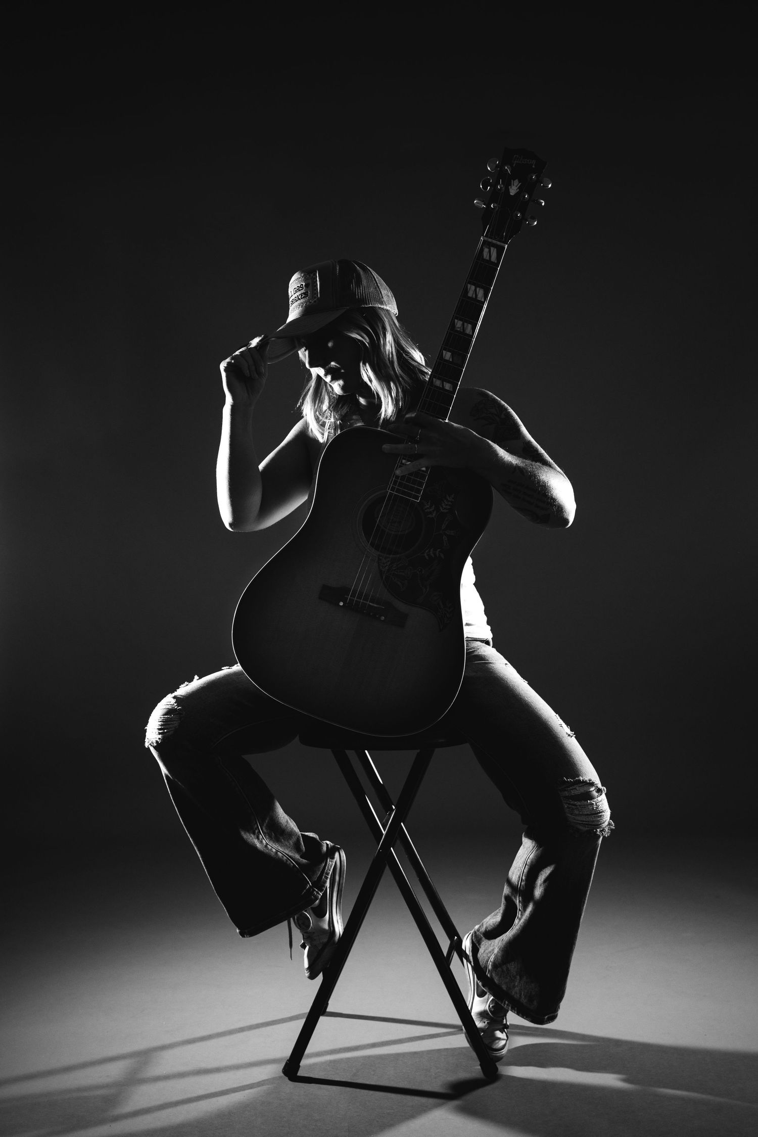 Silhouetted guitarist in cap sitting on stool with acoustic guitar in dramatic black and white lighting.