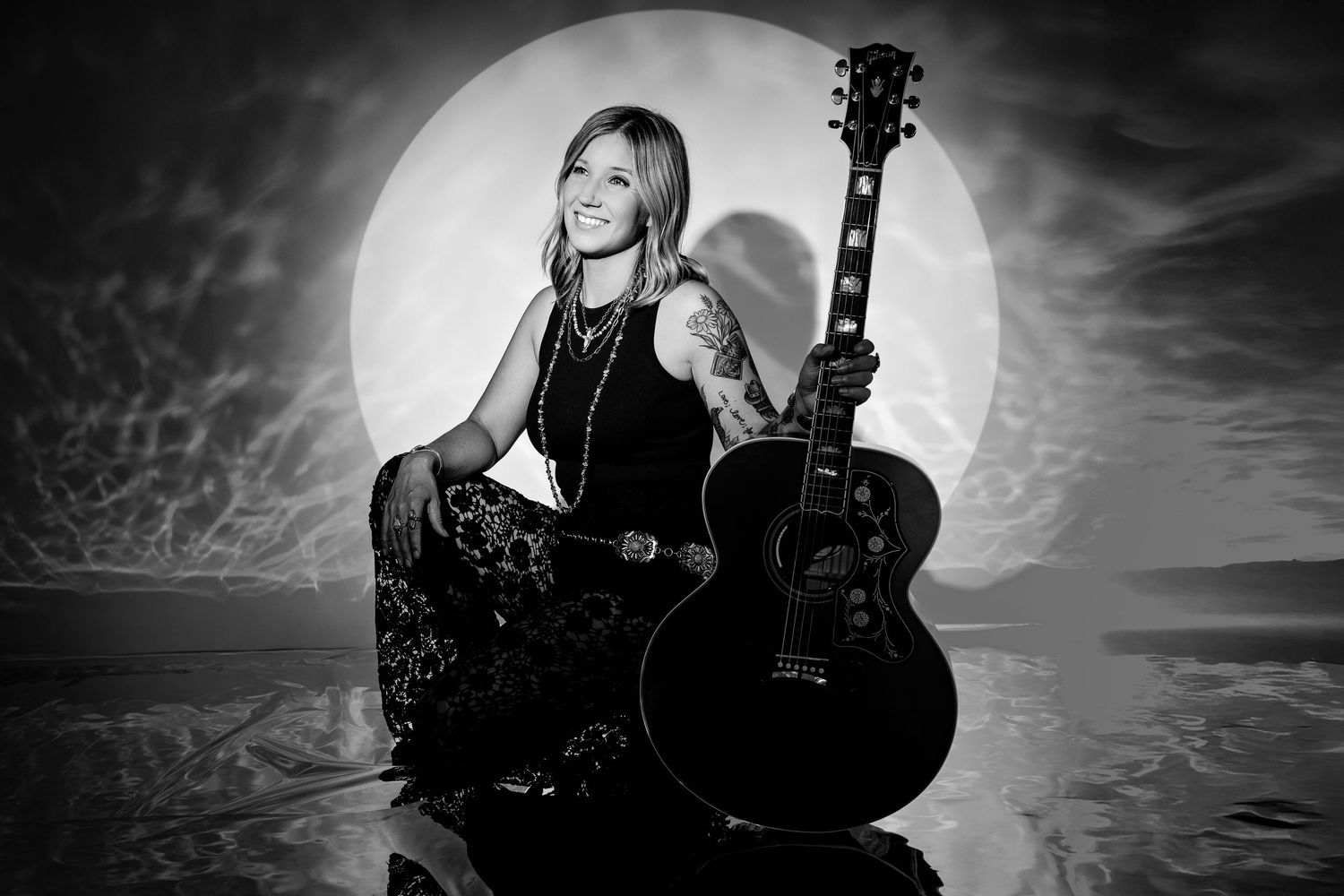 Black and white photo of tattooed woman holding Gibson guitar kneeling before large moon backdrop outdoors.