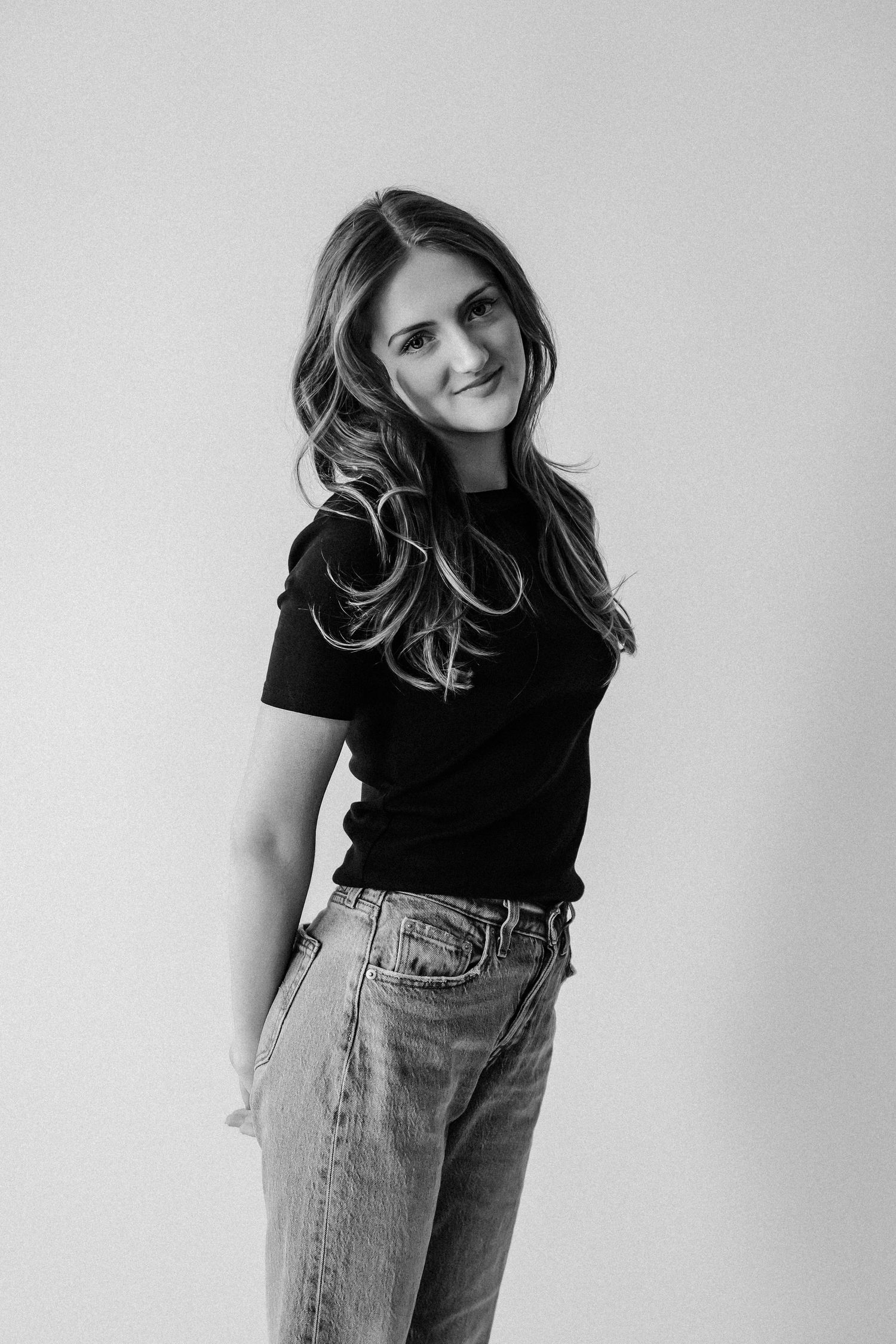 Young woman with long wavy hair smiling in black t-shirt and jeans in a black and white studio shot.
