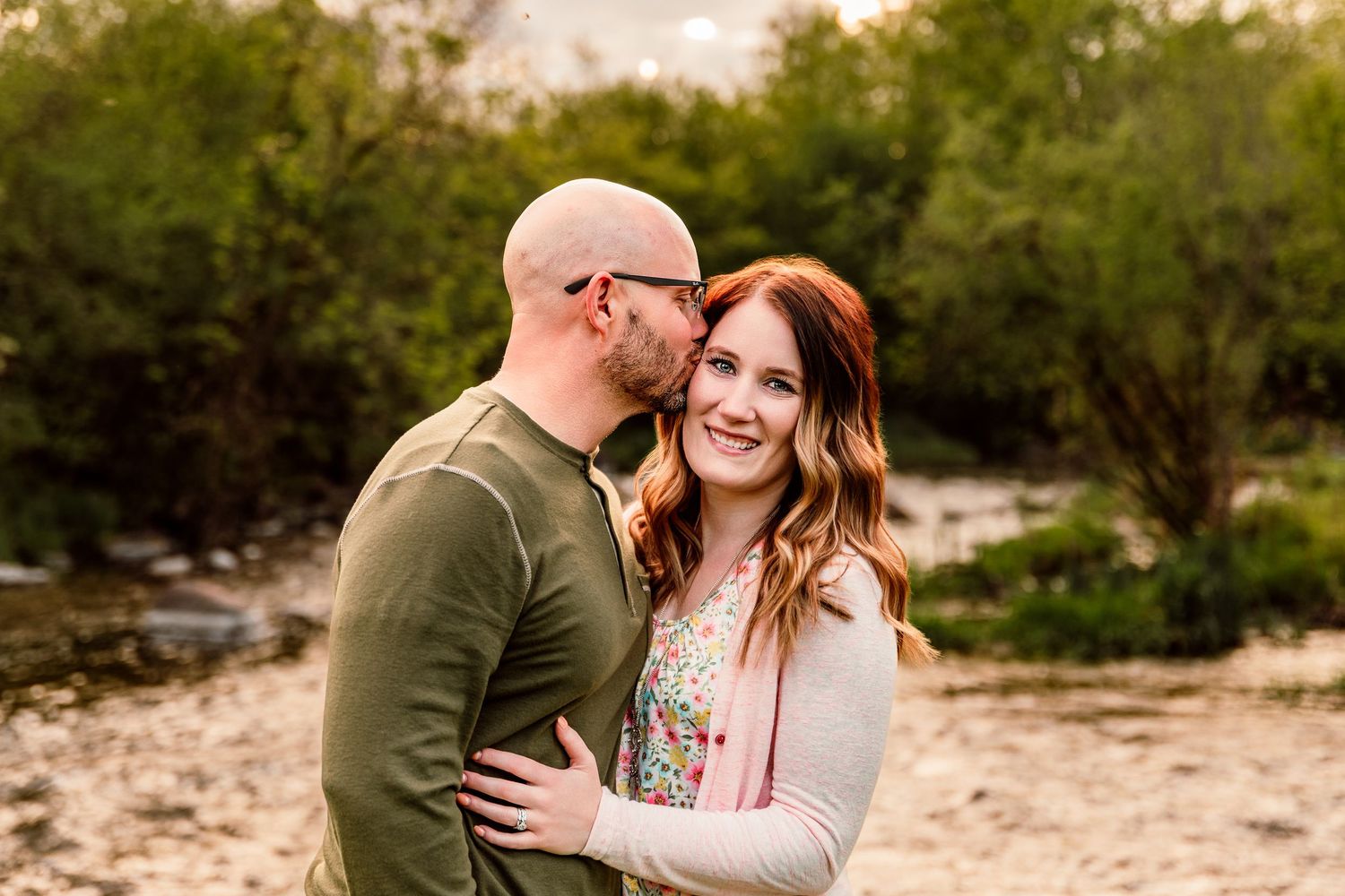 Couple embracing outdoors near a creek, woman smiling at camera while man kisses her cheek at golden hour.