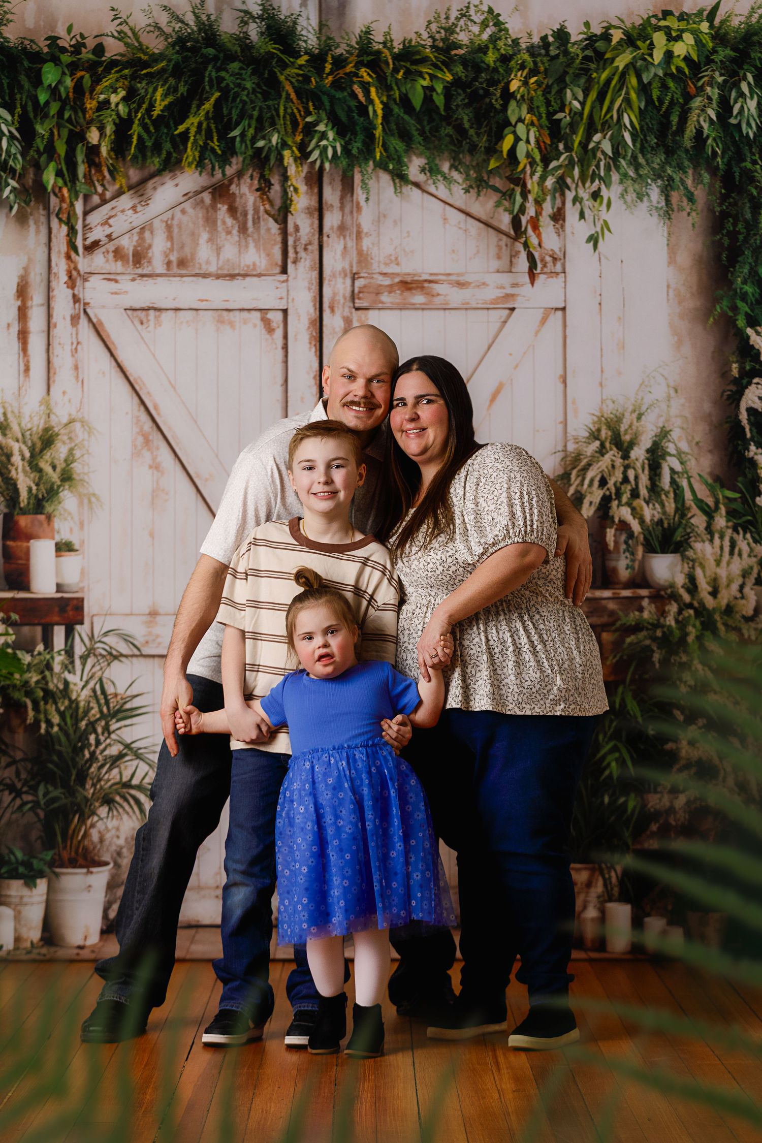 Family of four posing together in front of rustic barn doors with lush greenery backdrop on hardwood floors.