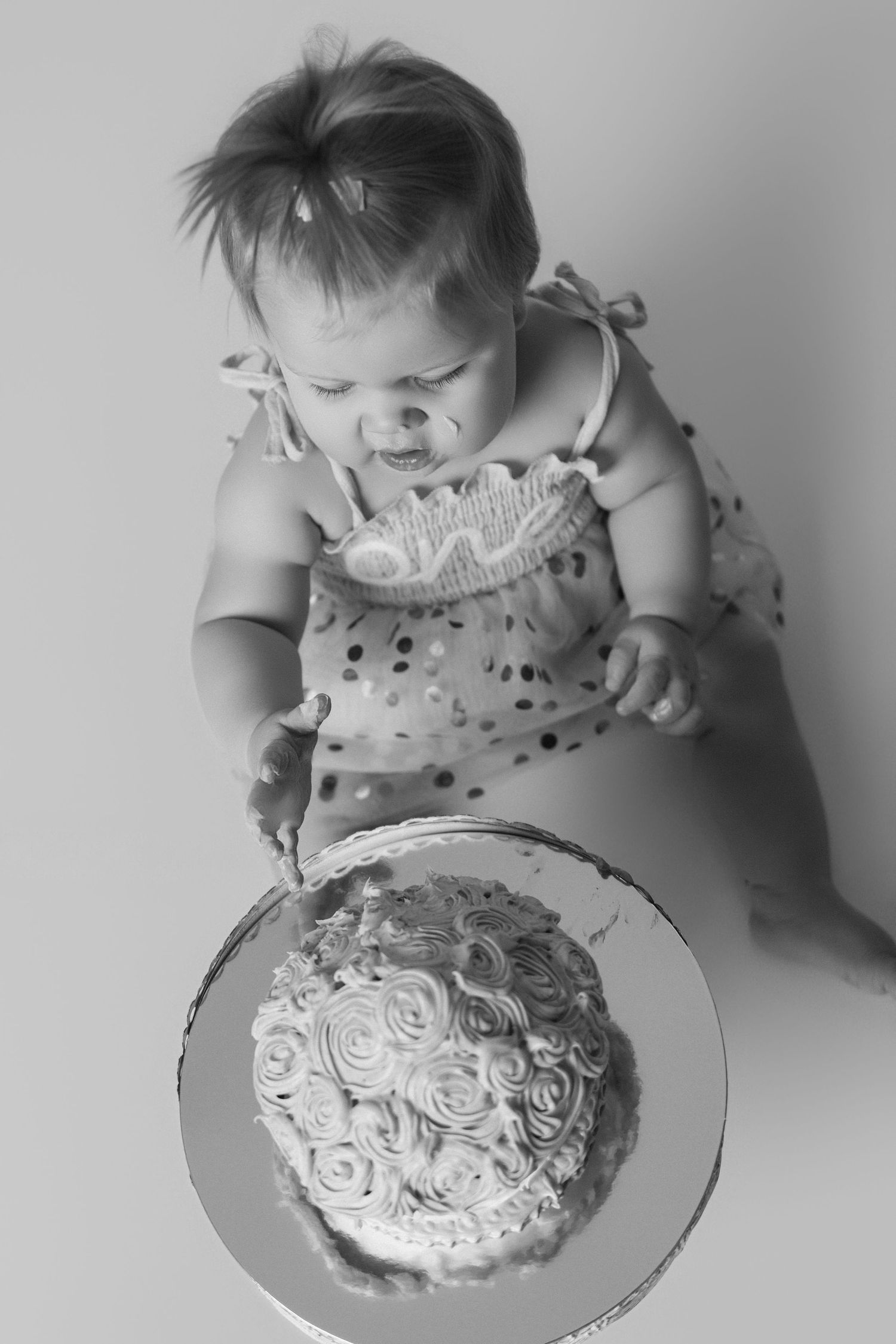 A baby in a polka dot dress leans over to examine a beautifully decorated white cake.