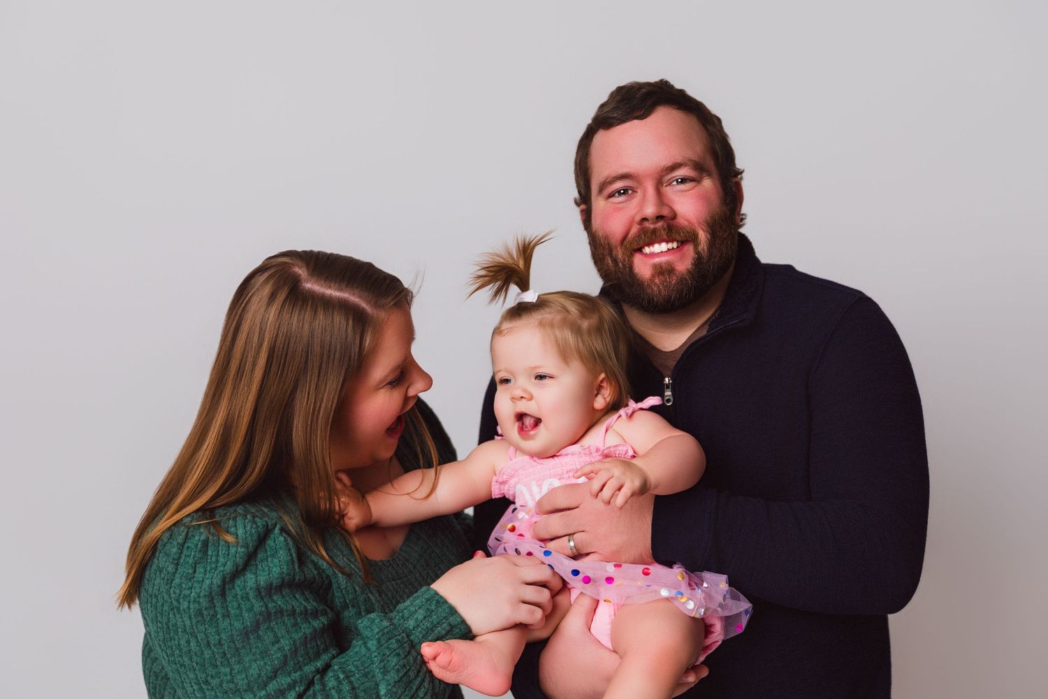 Family portrait with parents and laughing baby in pink dress against grey background.