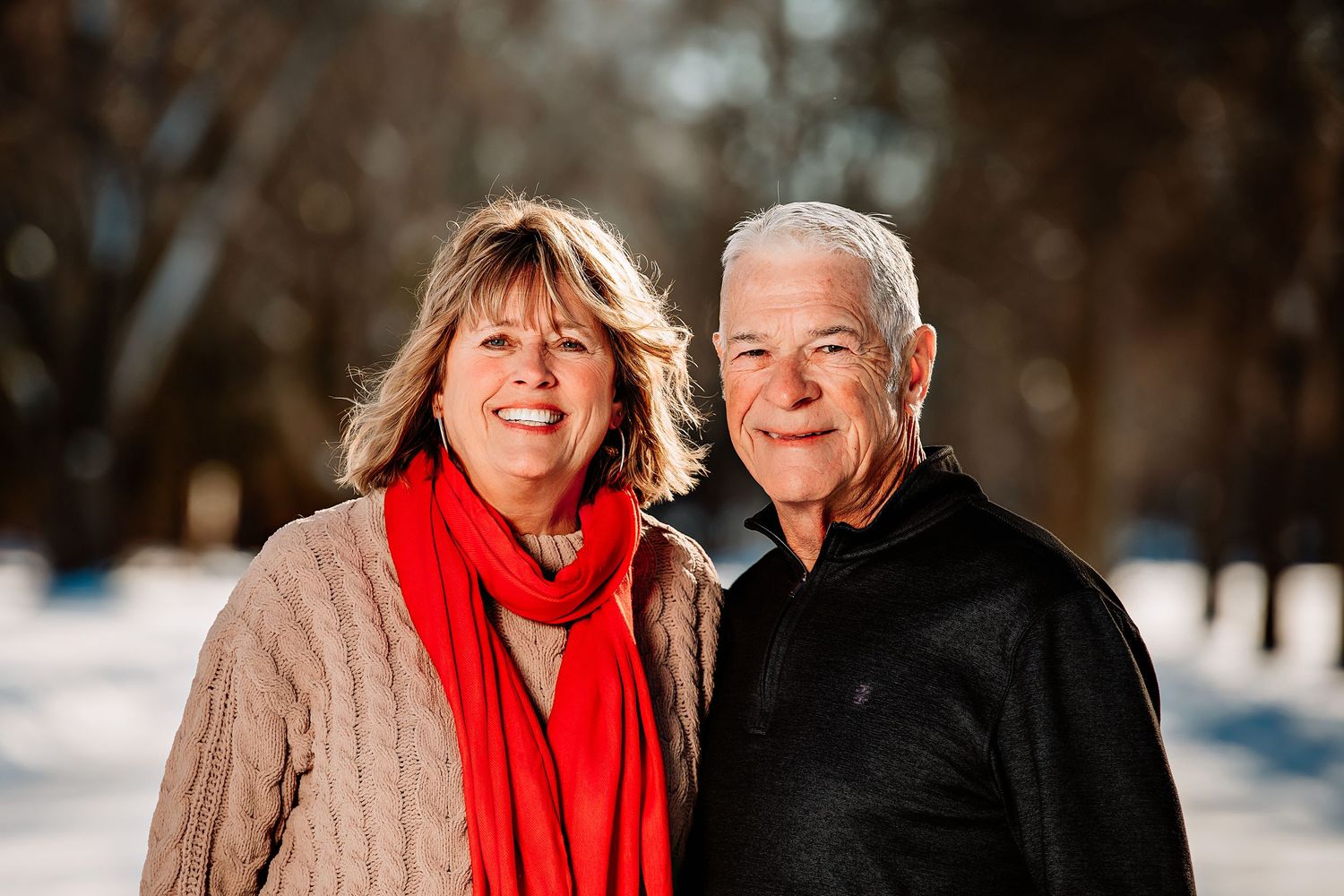 Two people wearing winter clothing smile together outdoors in a snowy setting with trees in the background.