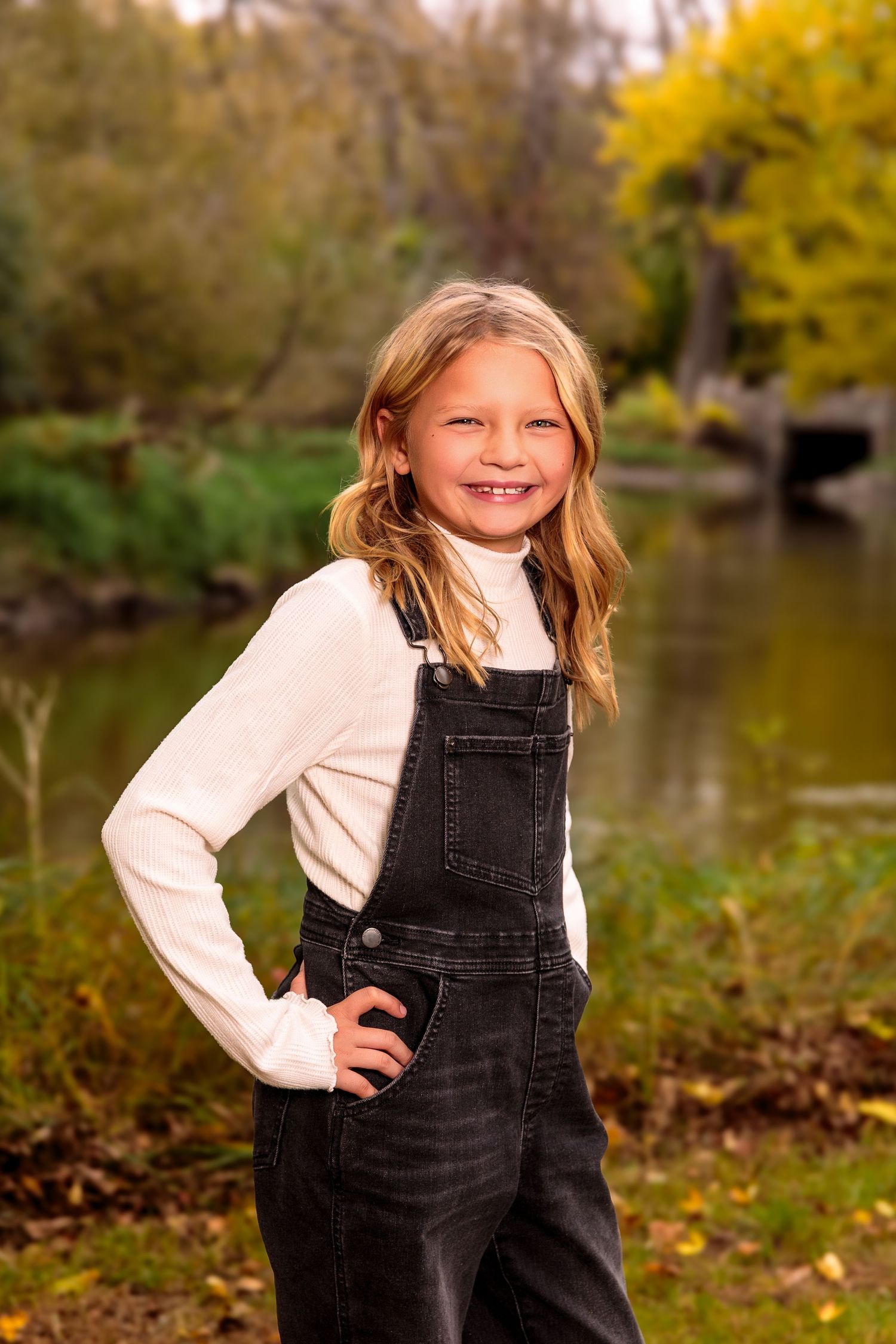 Young child in black overalls and white shirt plays outdoors during autumn near a pond.