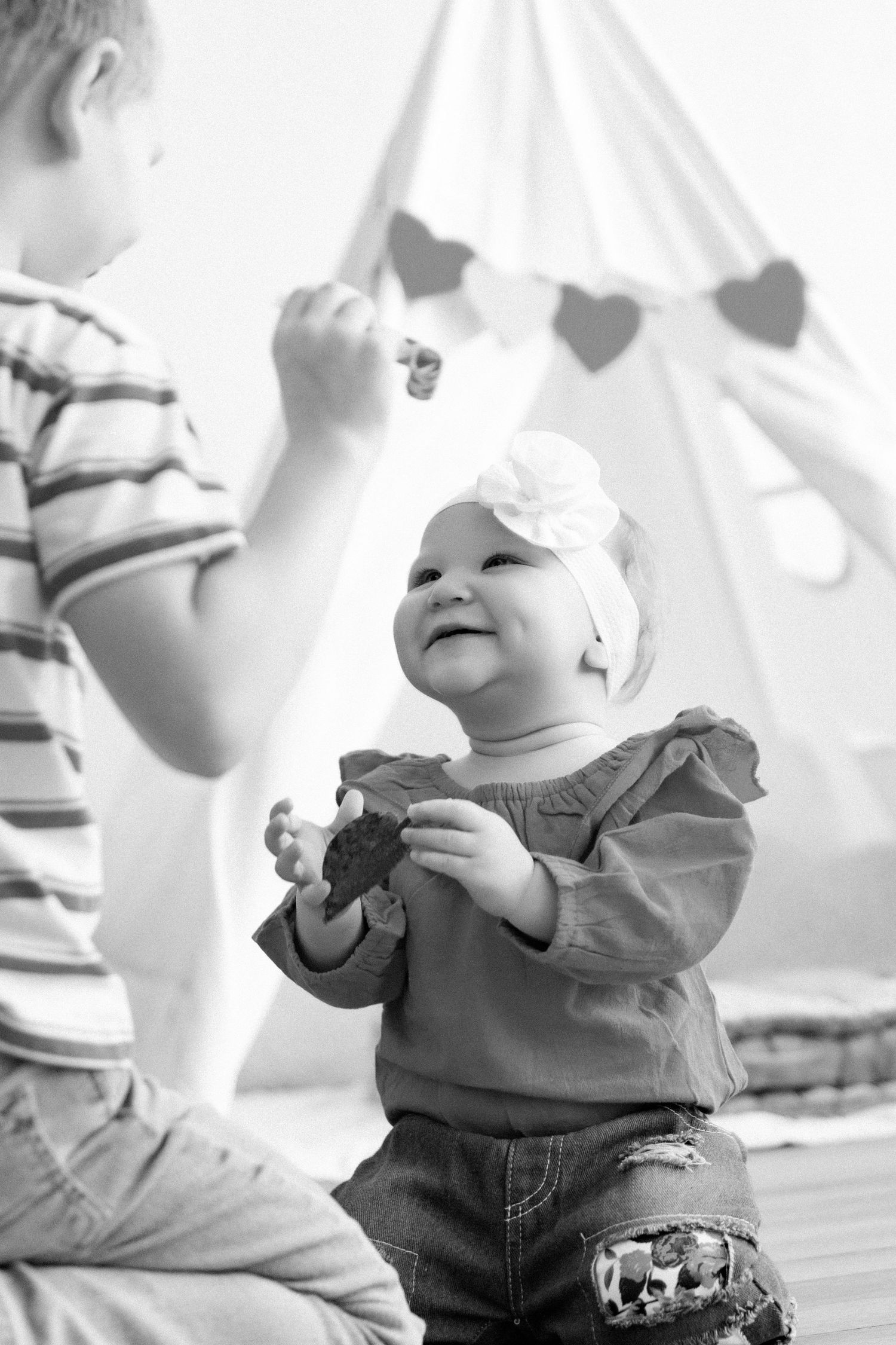 Black and white photo of baby playing with toys in front of teepee with heart garland.