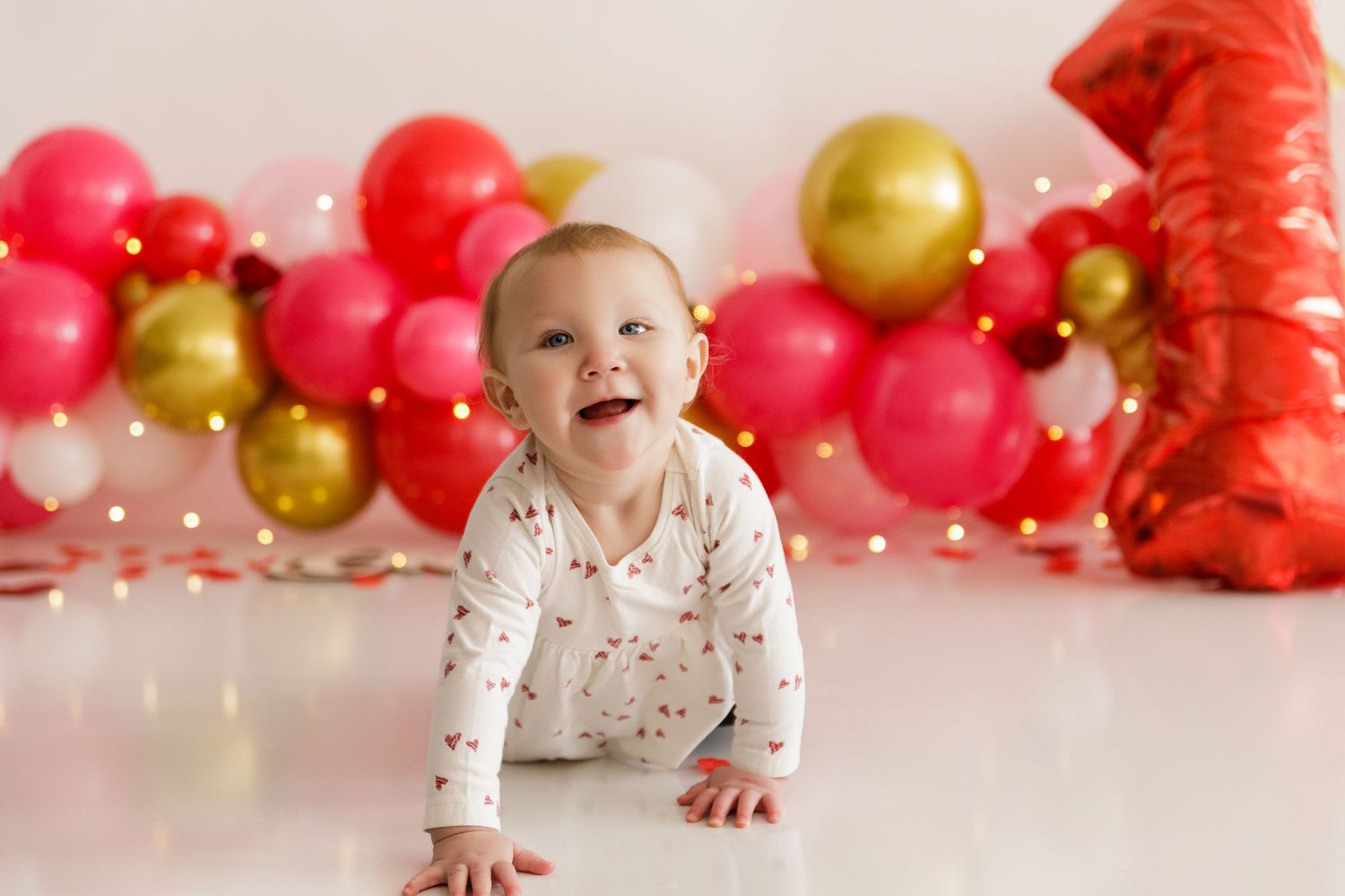 Baby in white outfit crawling on floor with red and gold balloons in background.