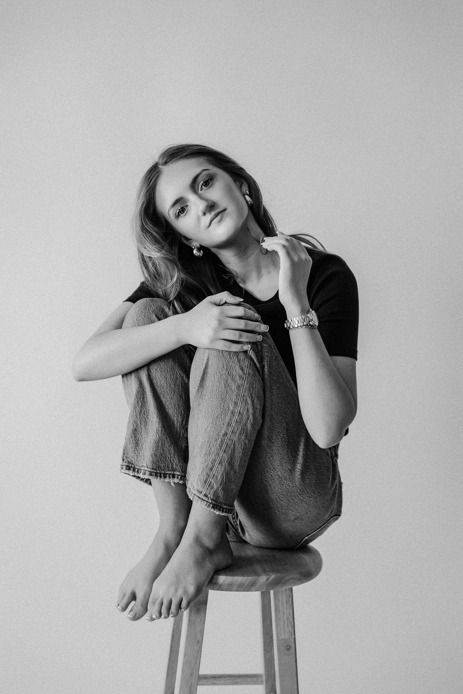 Black and white studio portrait of young woman seated on stool holding small flower, wearing black tee and jeans.