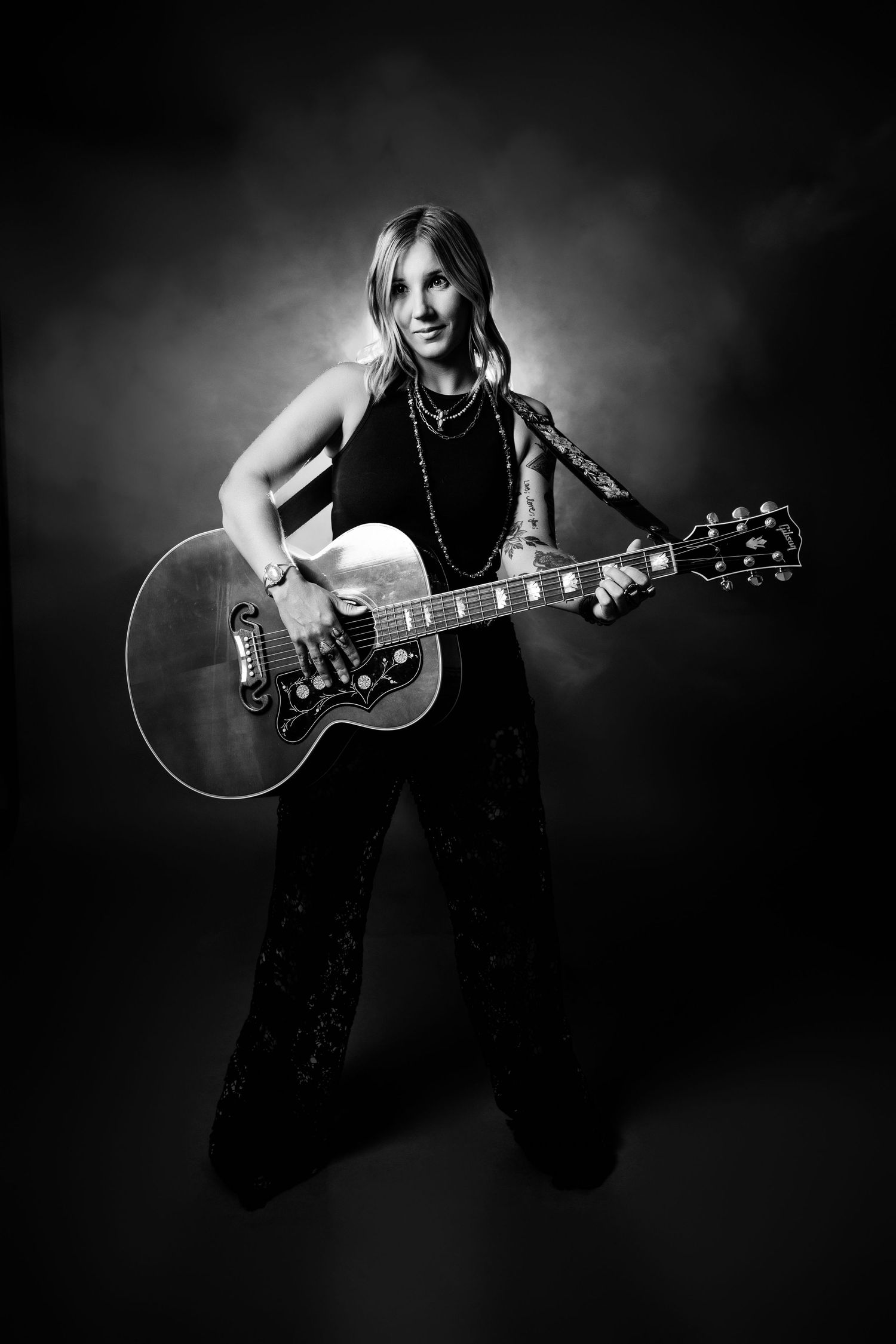 Black and white full-length portrait of female guitarist playing acoustic guitar in smoky studio.