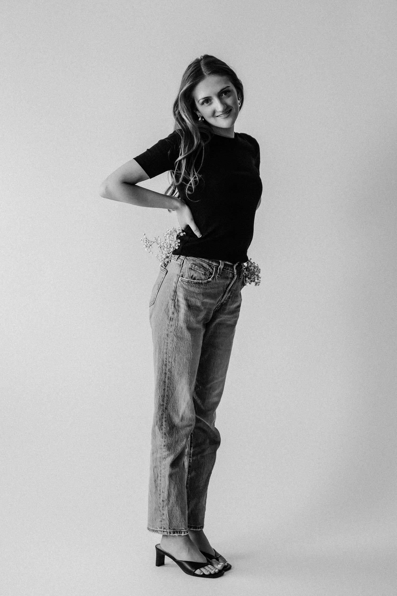 Smiling young woman in black tee and jeans holding flowers, posing in black and white studio photo.
