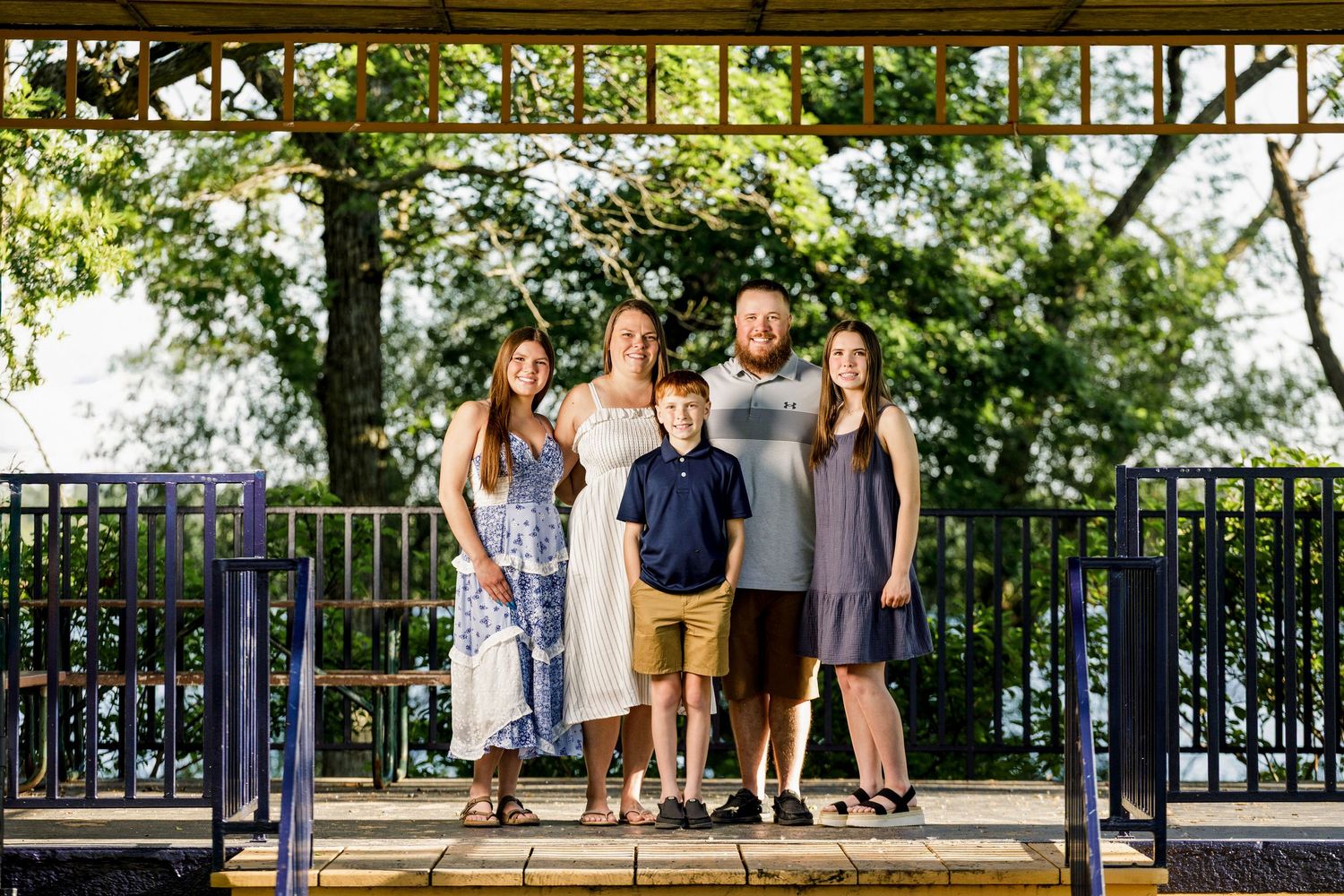 Family of five posing under a pergola outdoors, surrounded by lush green trees in a park setting.
