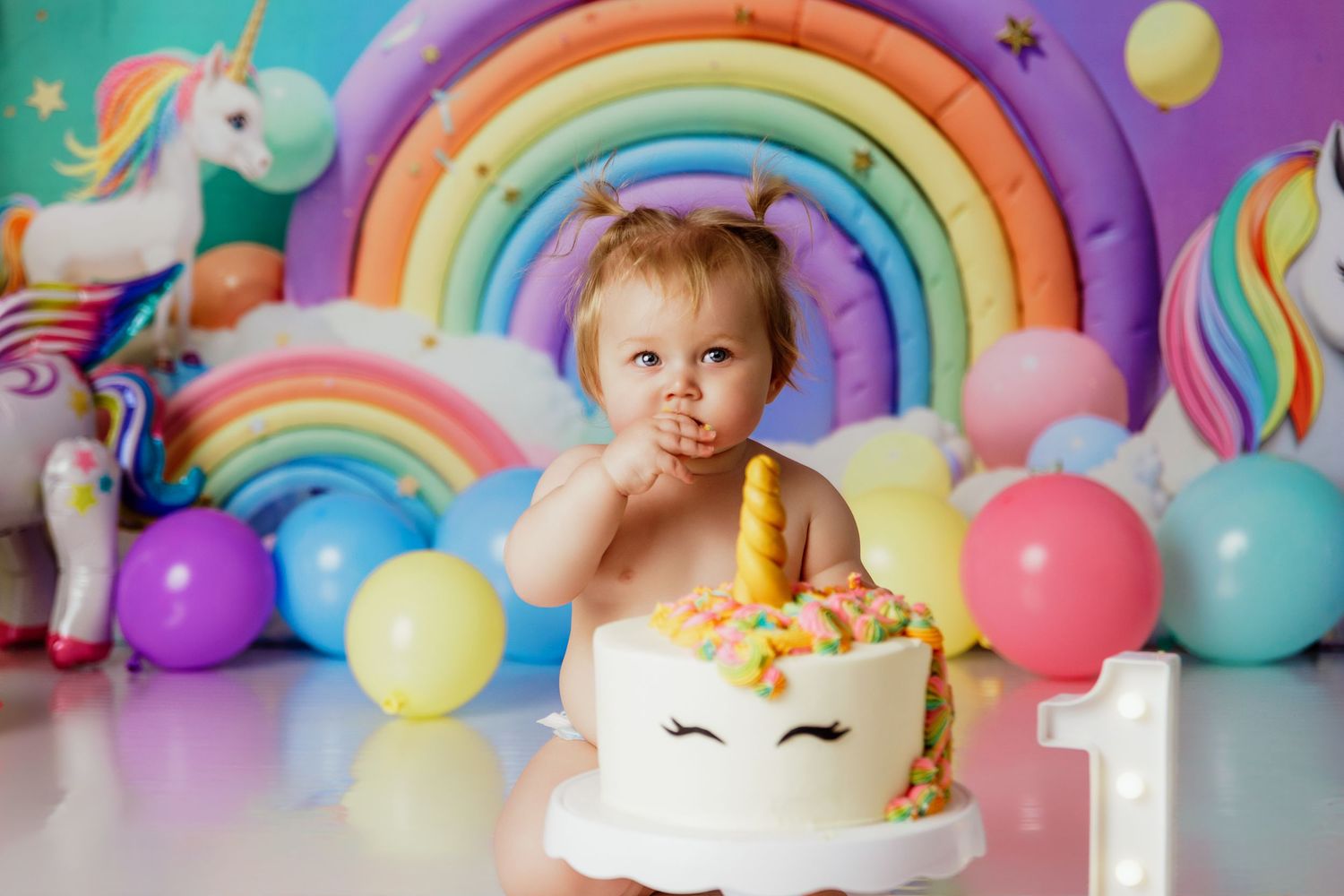 Baby girl eating unicorn birthday cake at colorful rainbow themed first birthday party.