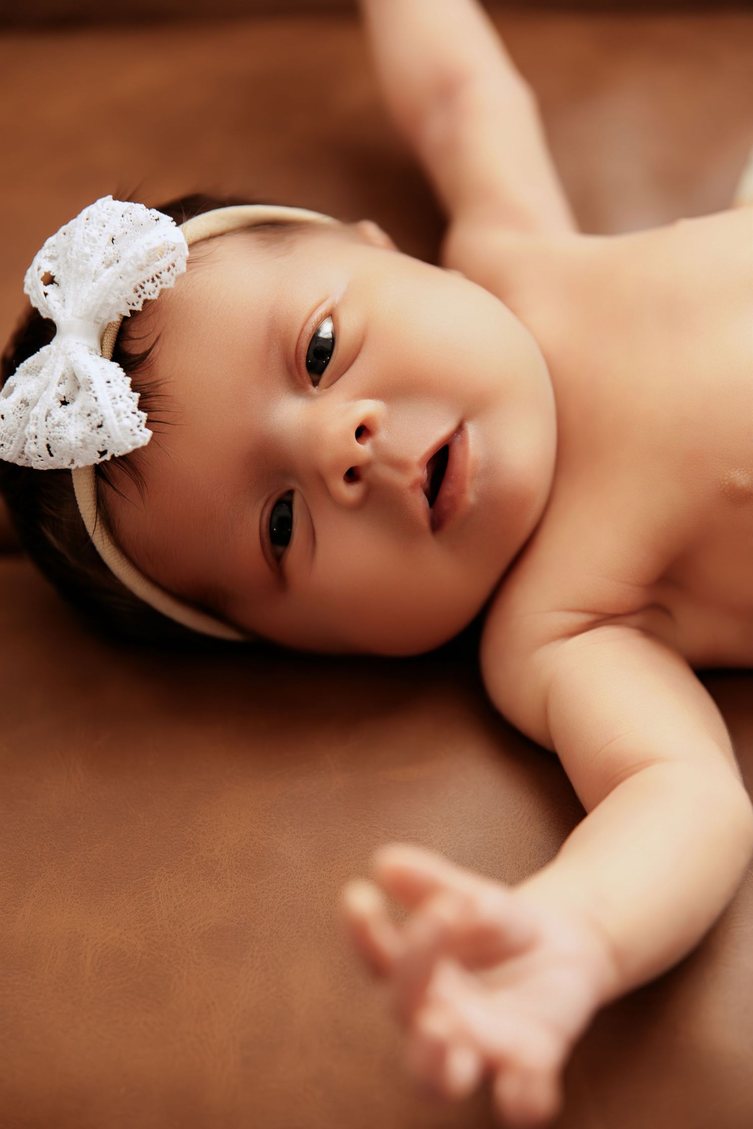 A sweet baby lies on a brown blanket wearing a delicate white lace headband.