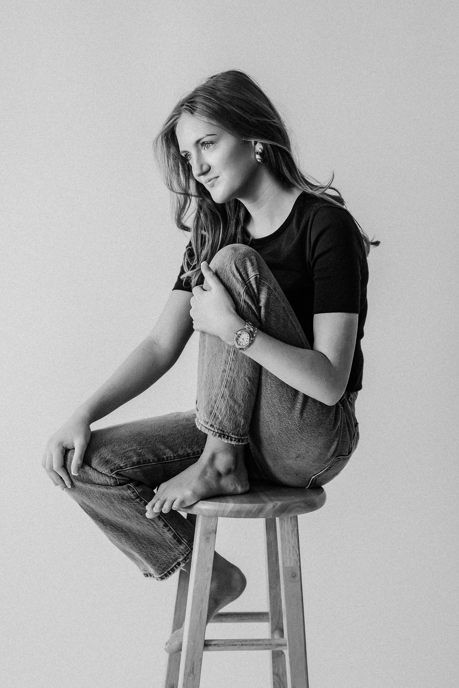 Black and white portrait of a young woman sitting on a wooden stool, wearing jeans and a black t-shirt.