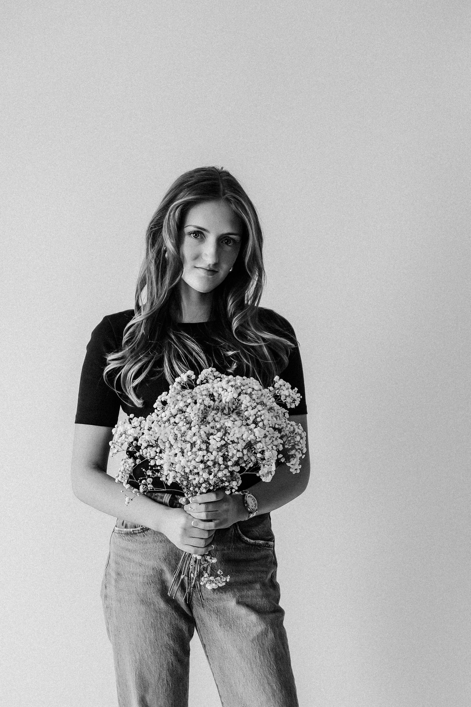 Black and white portrait of young woman holding bouquet of wildflowers, wearing black tee and jeans in studio.