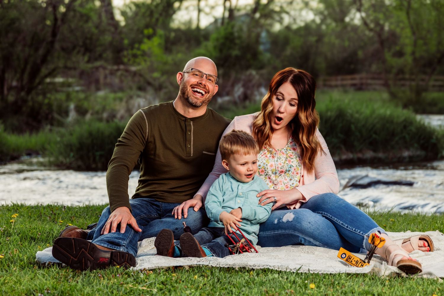 Happy family of three sitting on blanket outdoors, laughing and enjoying nature by a river.