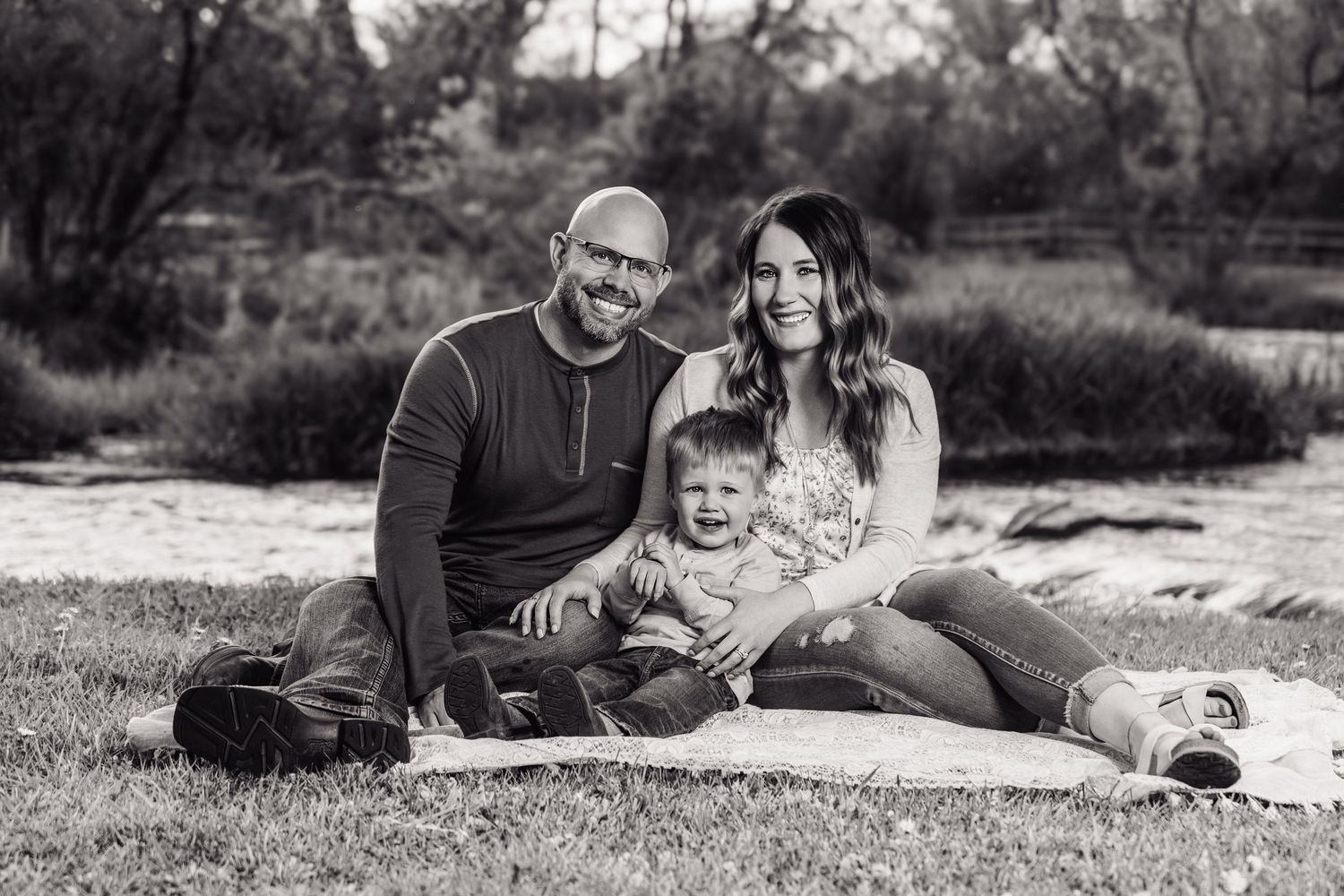 Black and white family portrait of parents and toddler sitting on blanket outdoors near a river.